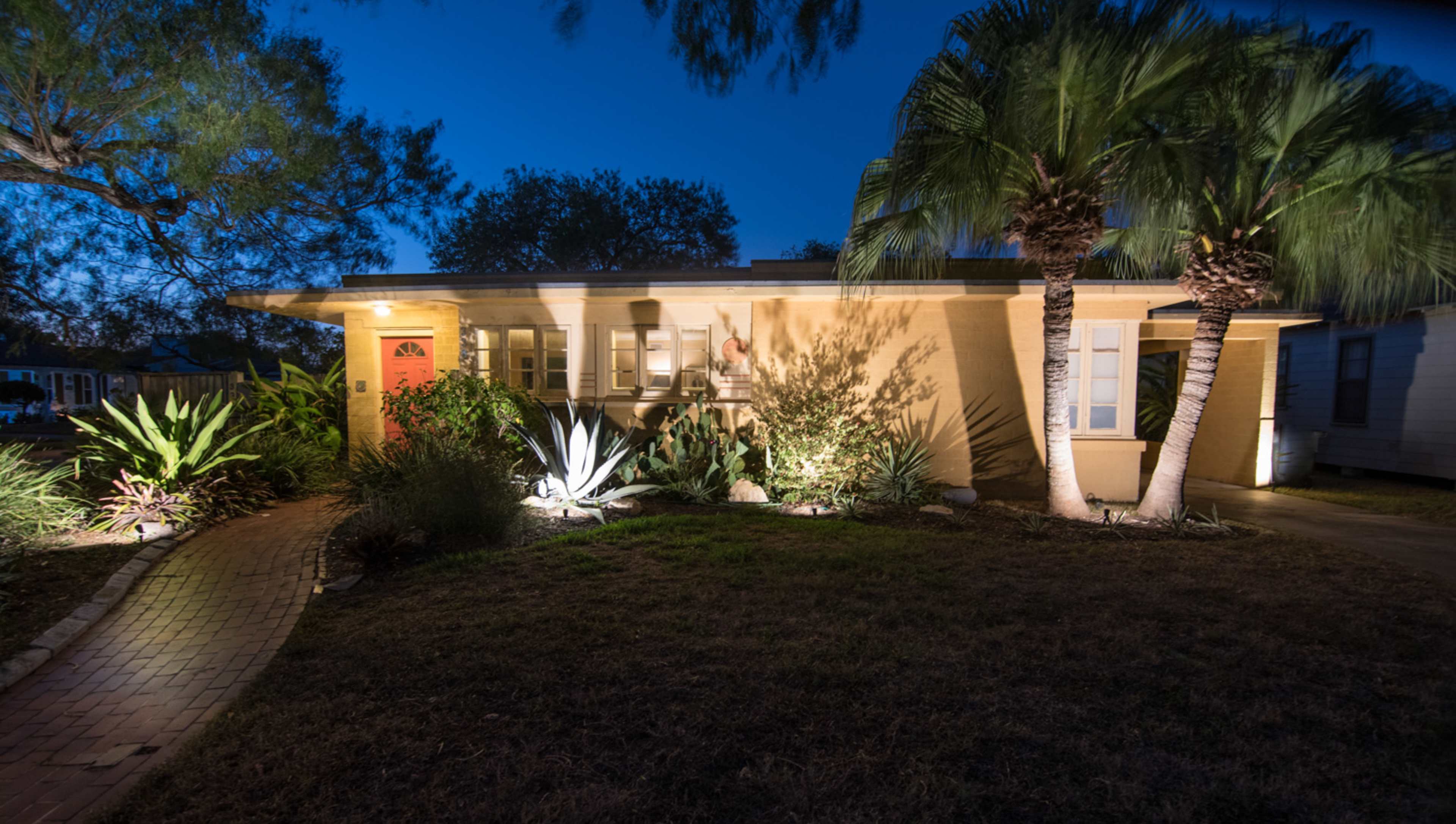 A yellow house is illuminated at night, surrounded by various plants and palm trees, with a brick pathway leading to the front door.