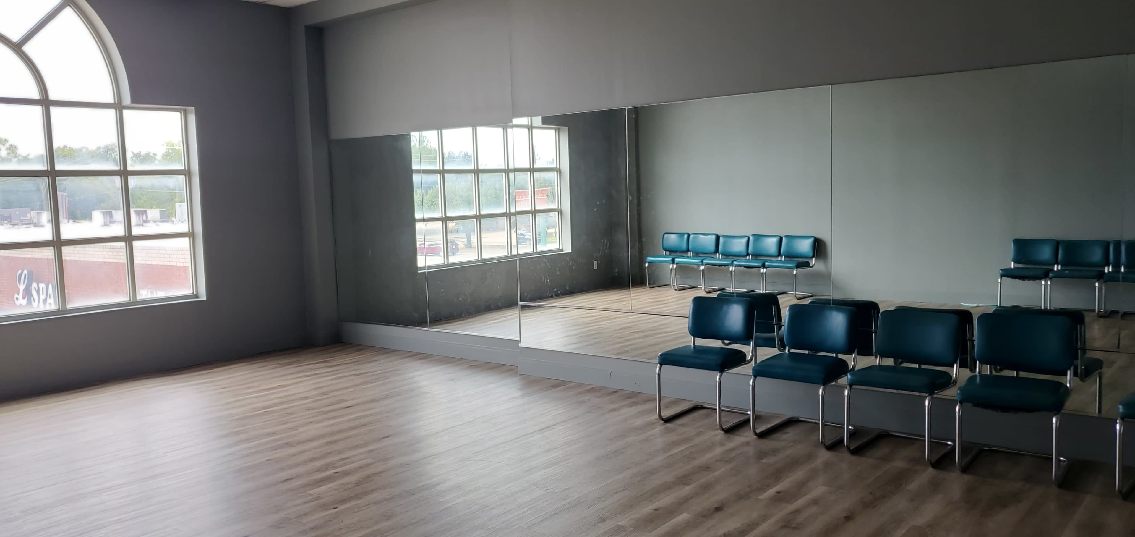 An empty room with wooden floors, a large mirrored wall, and a row of blue chairs arranged against the opposite wall.