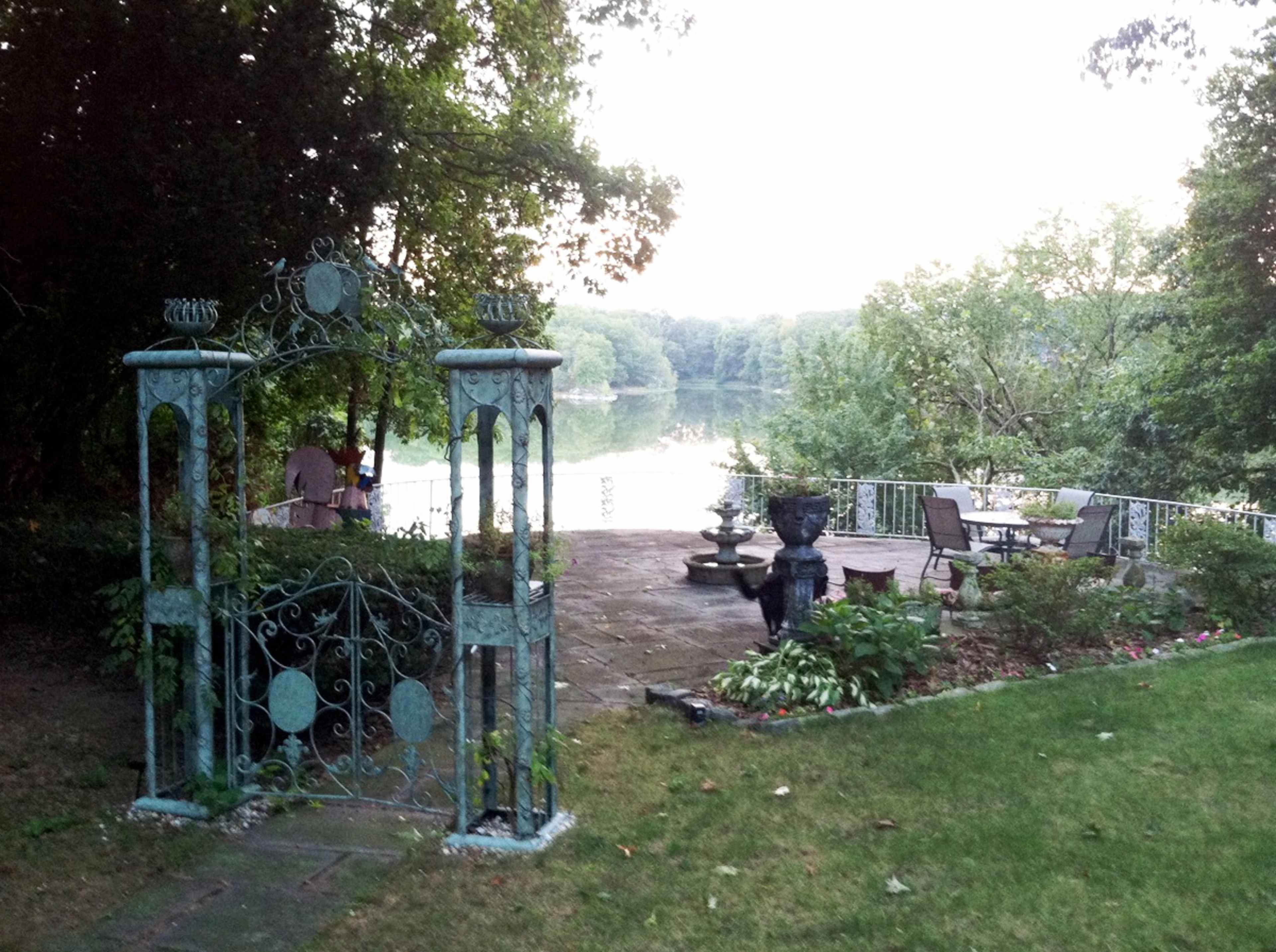 A wrought iron gate opens to a stone patio overlooking a calm lake, surrounded by trees and garden plants.