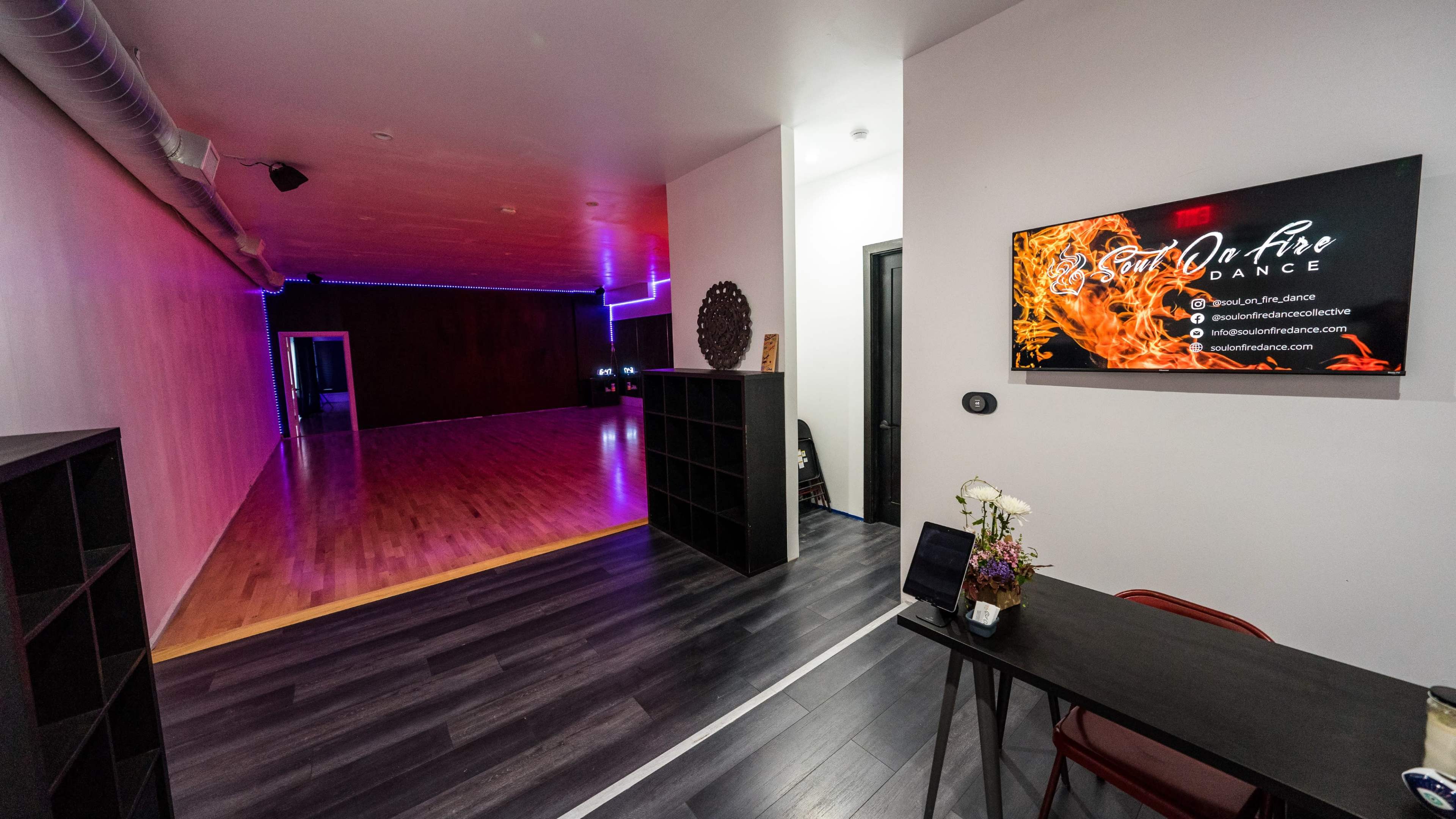 A modern dance studio featuring a spacious wooden floor, illuminated with colored lights, and a reception area with a decorative table and a digital display.