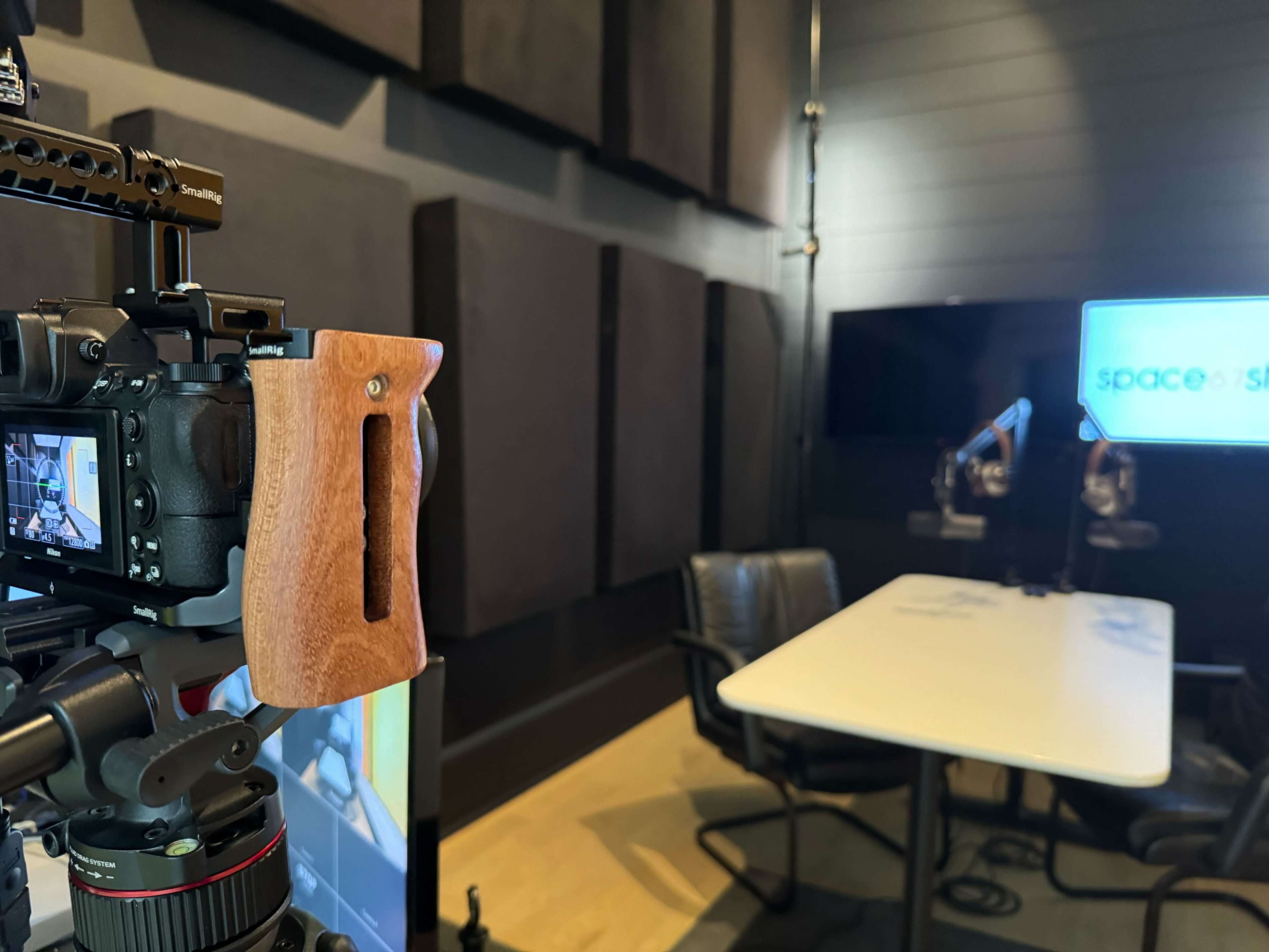 A camera mounted on a tripod, focused toward a prepared studio space with two chairs and a table, featuring soundproof panels on the walls.