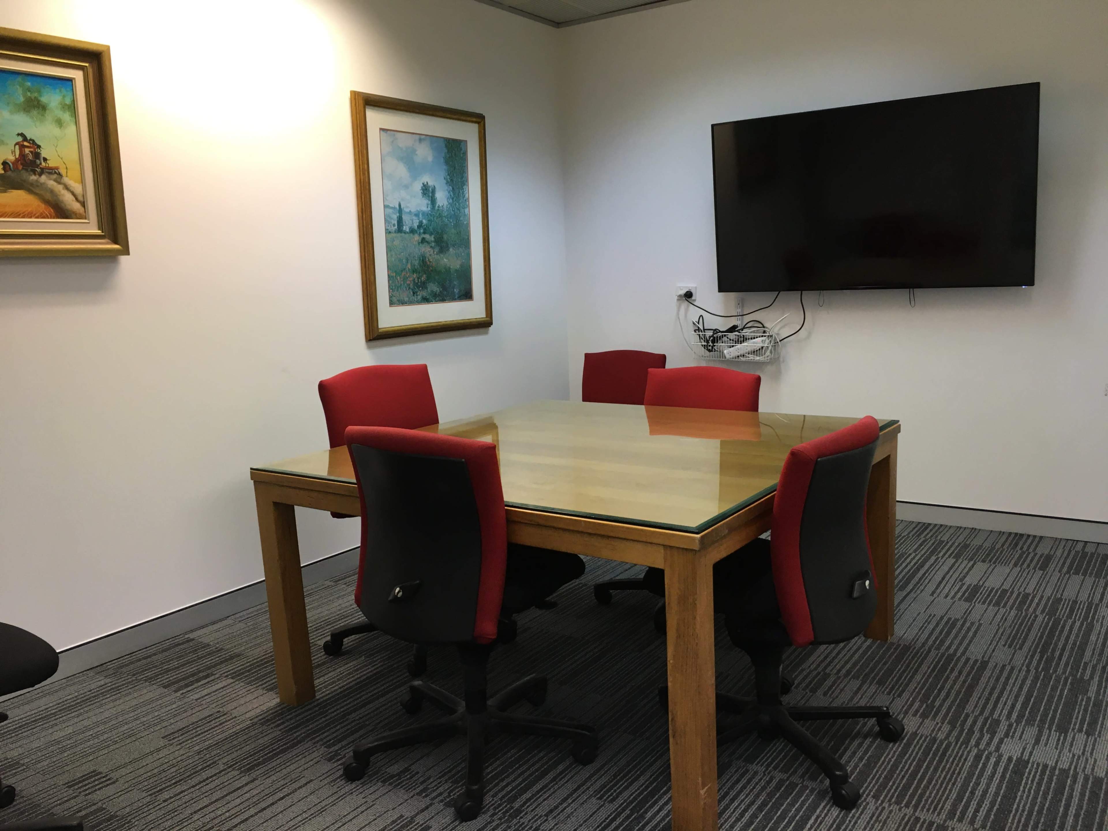 A conference room features a glass-top table surrounded by four red chairs, with two framed paintings on the walls and a mounted television.