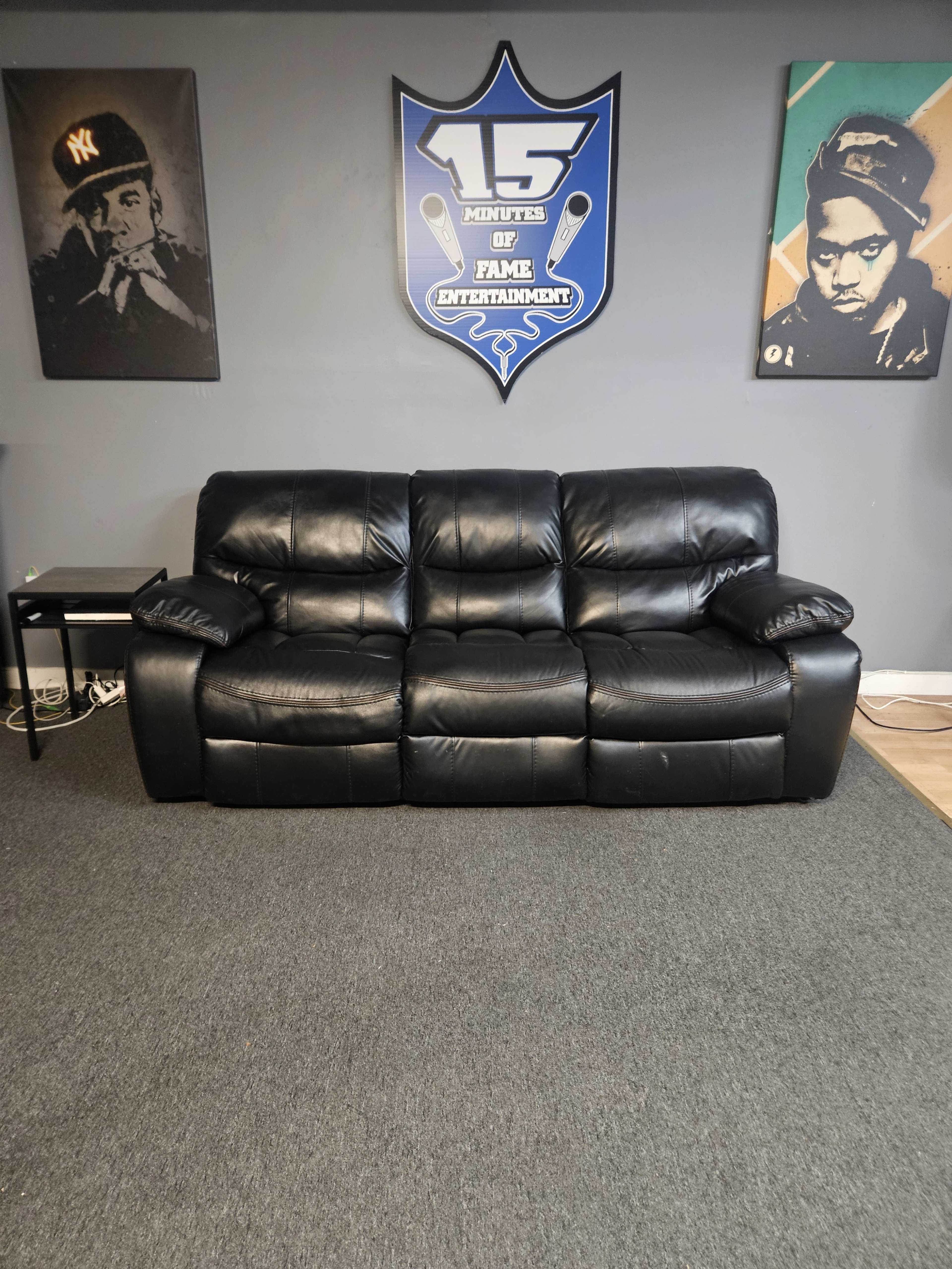 The image shows a black leather sofa positioned against a wall adorned with artwork and a shield-shaped sign that reads "15 MINUTES OF FAME ENTERTAINMENT."