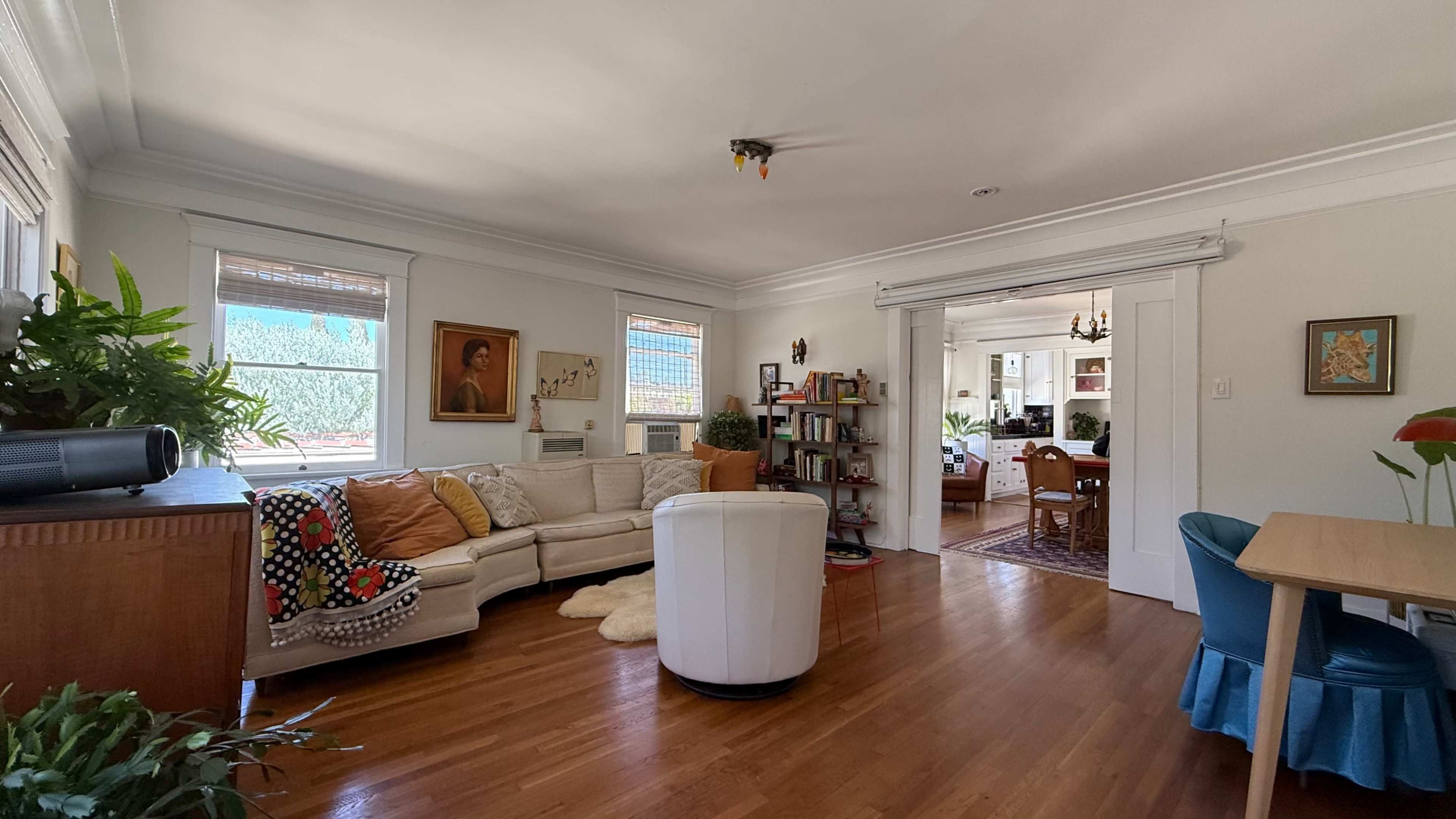 The image shows a bright living room with a beige sectional sofa, wooden flooring, a round white ottoman, and a doorway leading to another room.