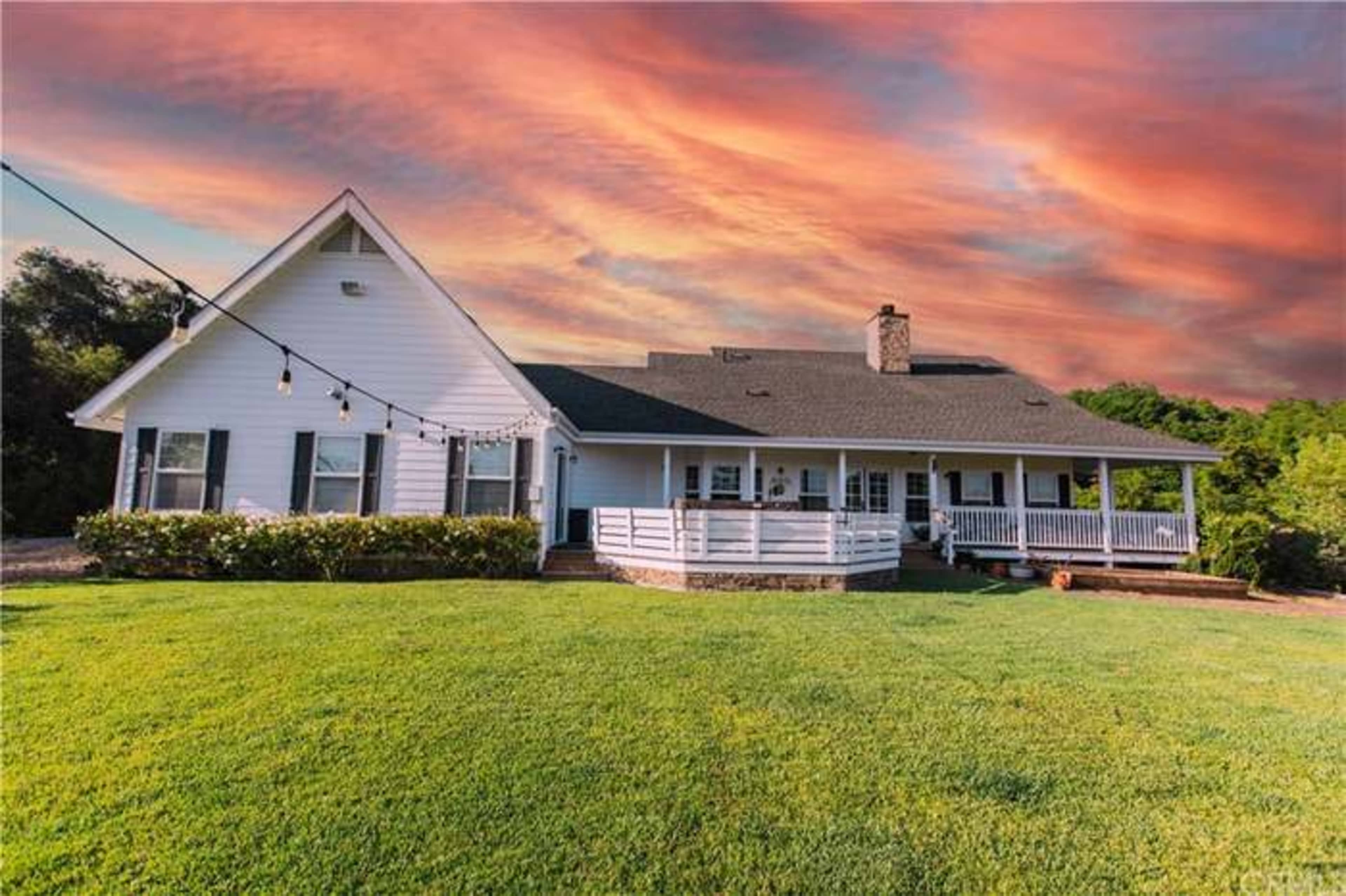 A white house with a large porch is set against a colorful sunset sky, surrounded by green lawn and landscaping.
