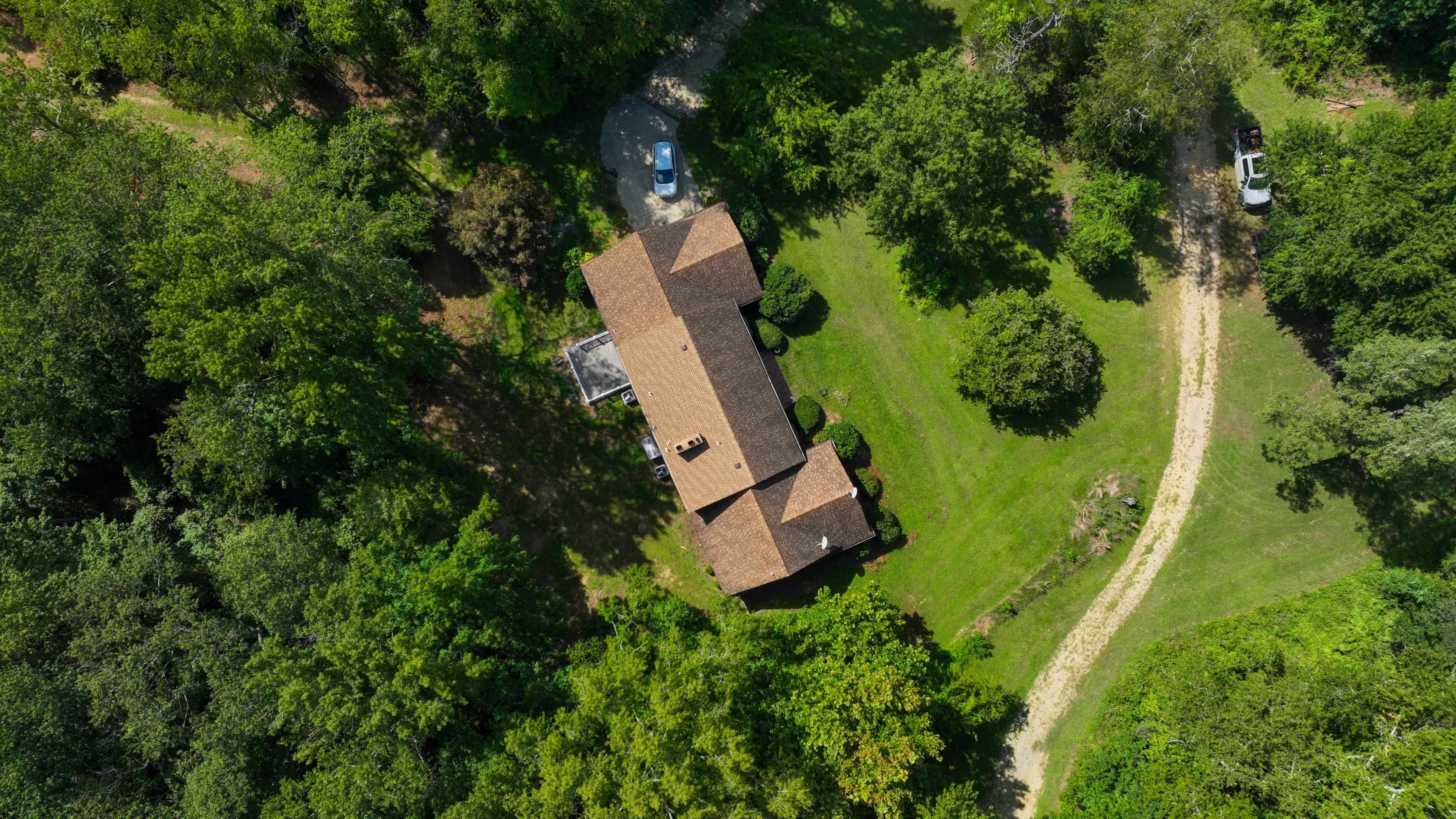 The image shows a house surrounded by greenery, with a winding dirt path leading to it and several vehicles parked nearby.