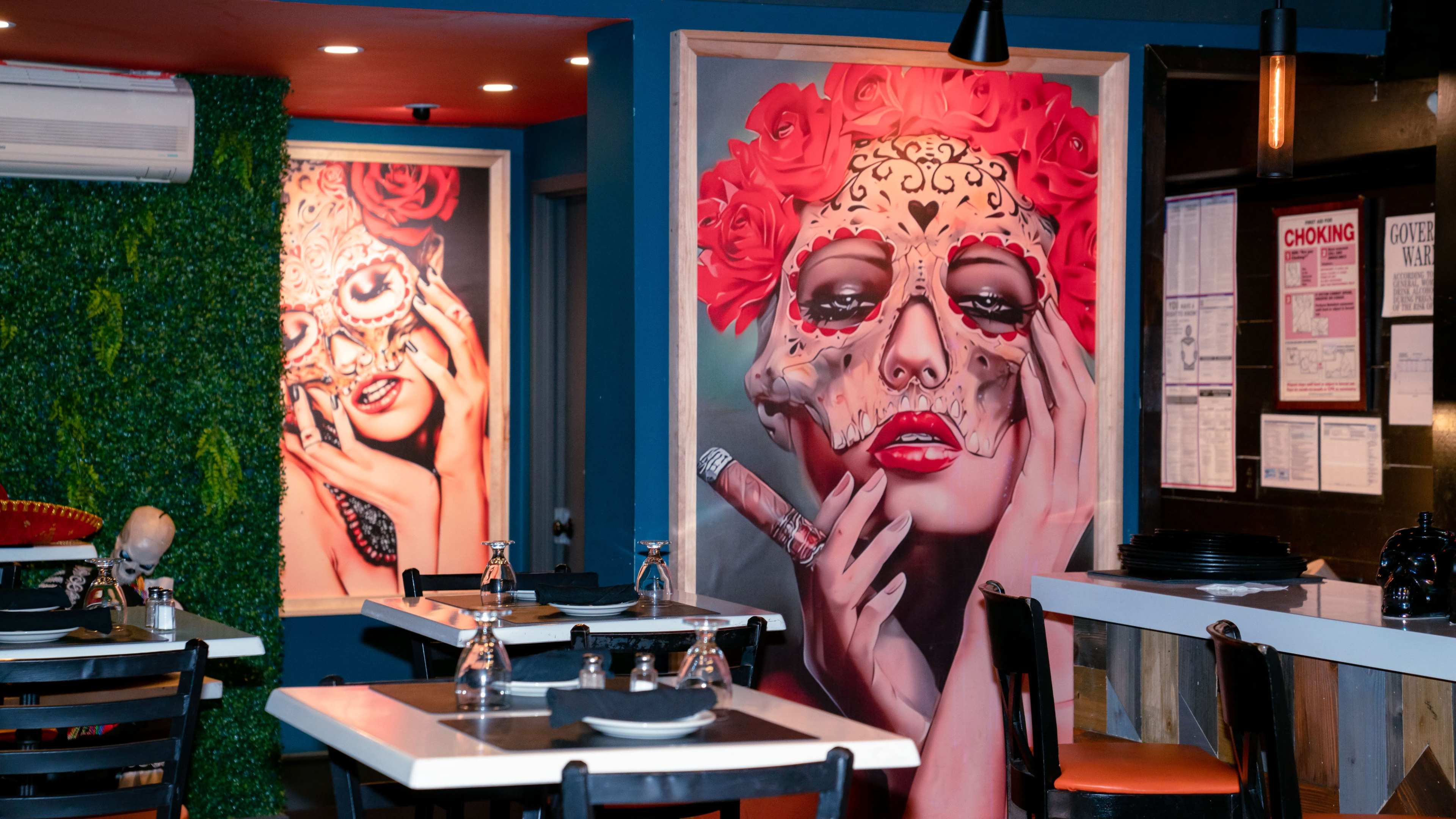 The image shows a vibrant restaurant interior featuring two large, colorful portraits of a woman with sugar skull makeup, surrounded by decor elements like greenery and dark furniture.