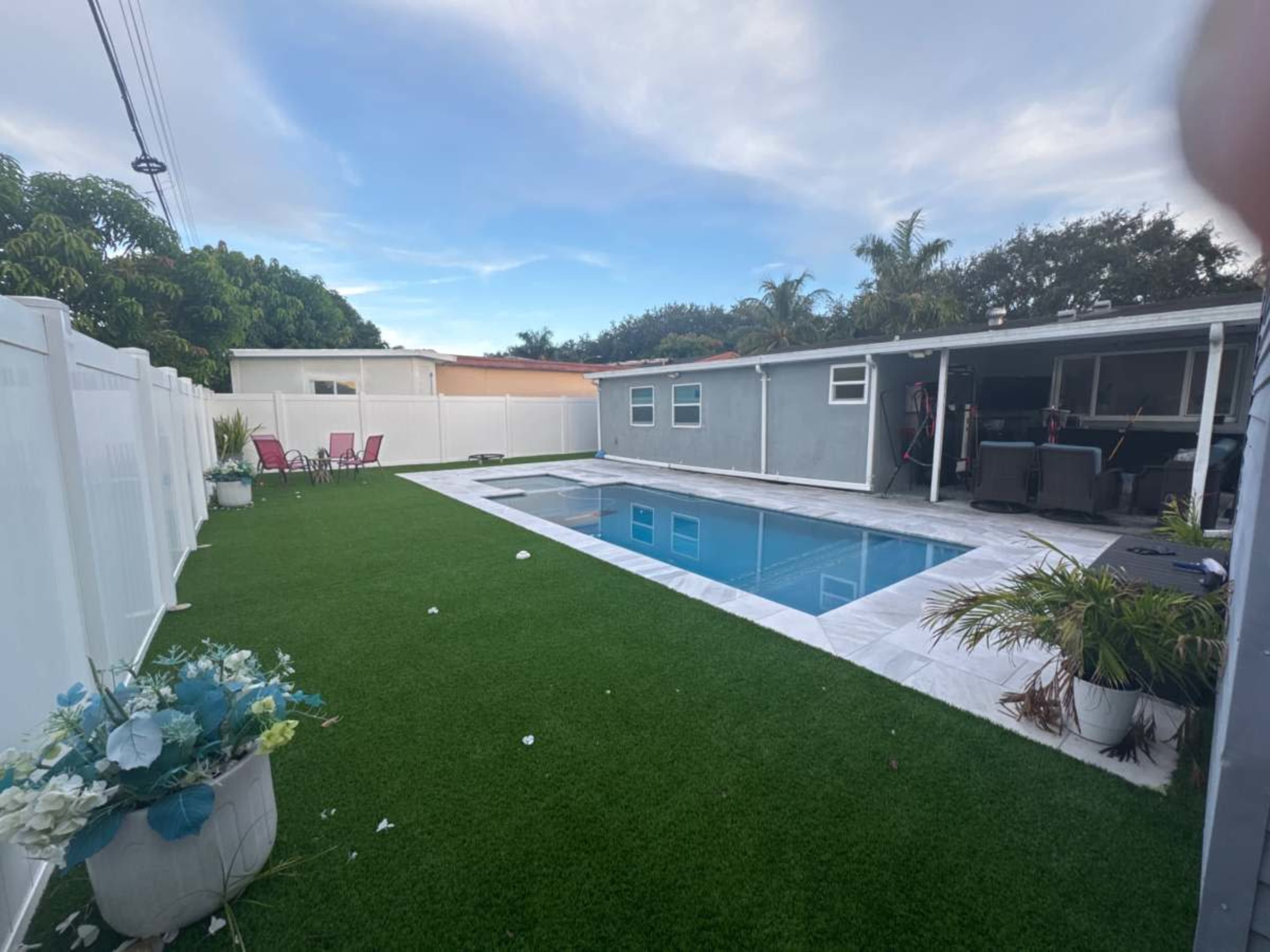 The image shows a backyard with a swimming pool surrounded by artificial grass, a patio area, and several potted plants.