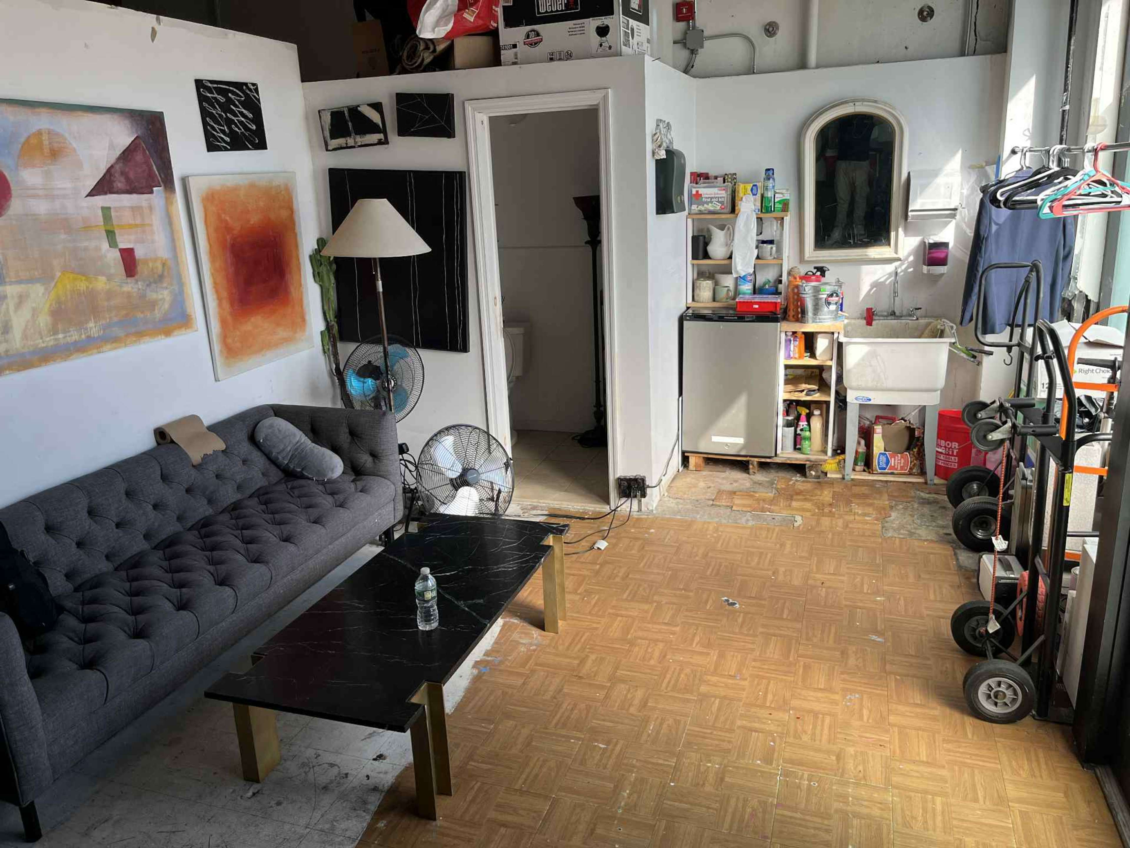 The image shows a small living area with a gray tufted sofa, a black coffee table, and a kitchenette area filled with various items.