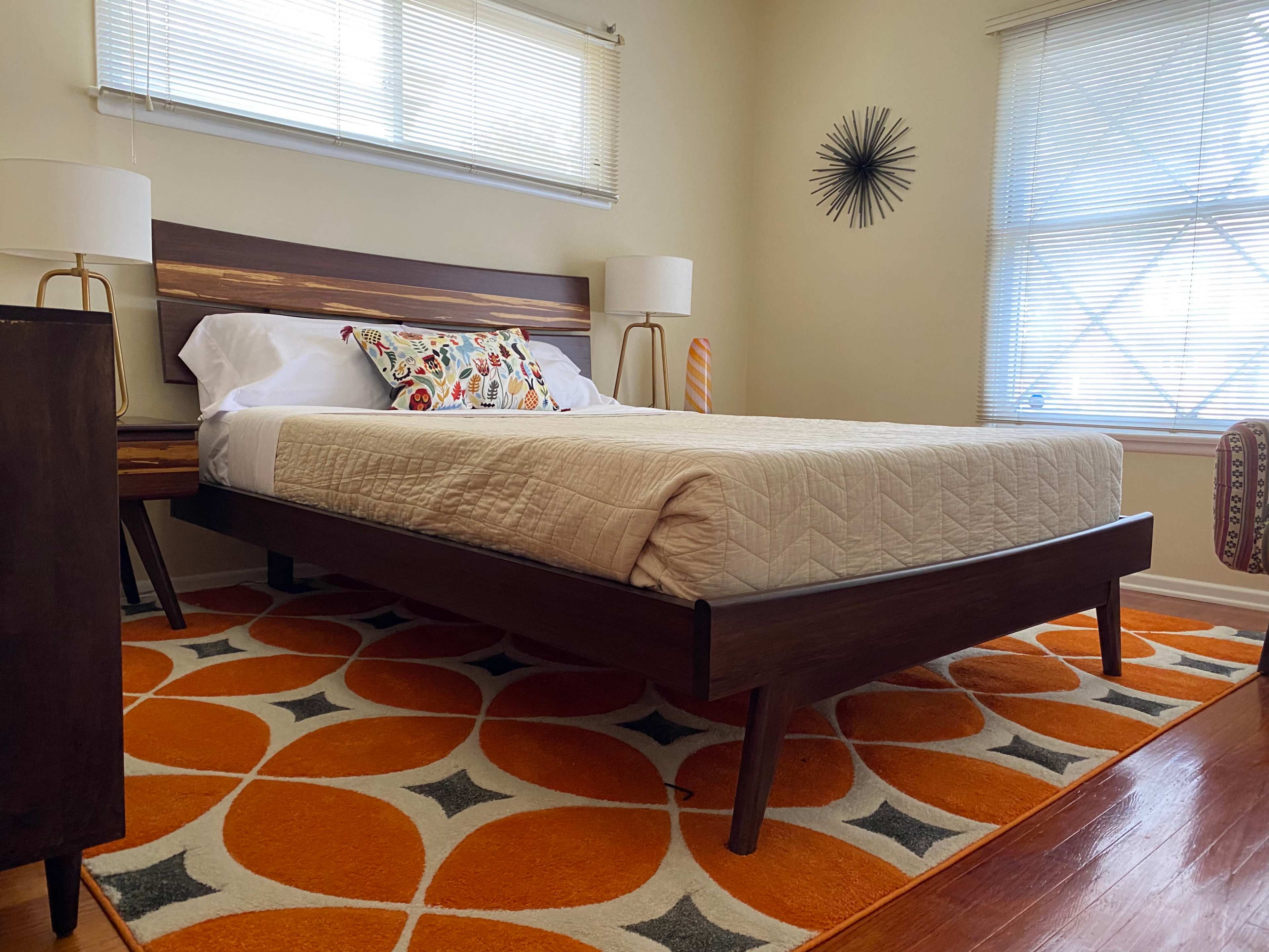 A bedroom featuring a wooden bed with a beige quilt, two bedside lamps, and an orange and white patterned rug on hardwood flooring.