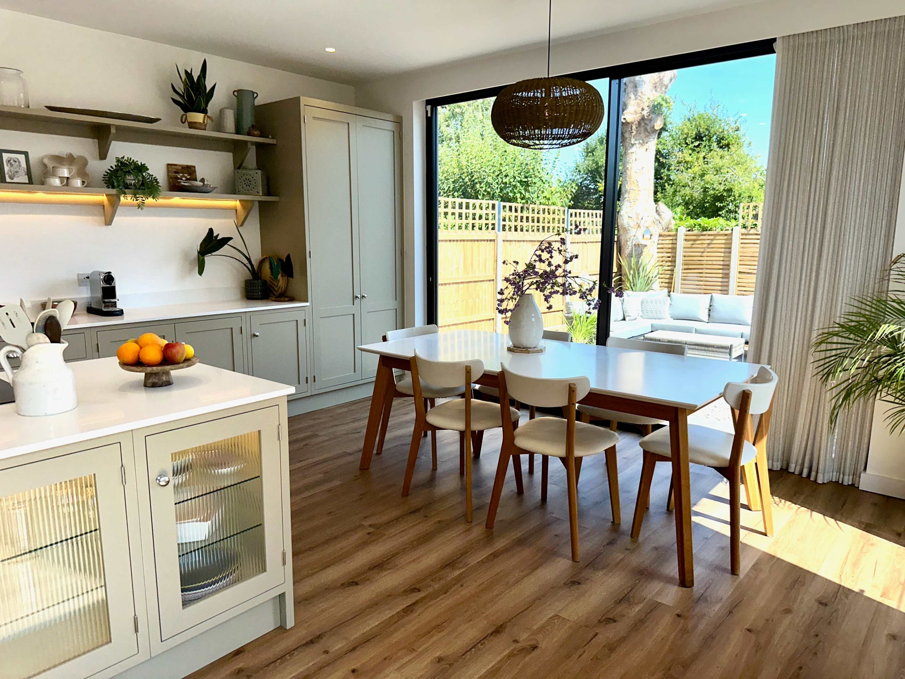 A bright dining area features a wooden table surrounded by chairs, with large windows overlooking a garden and a cozy seating area.