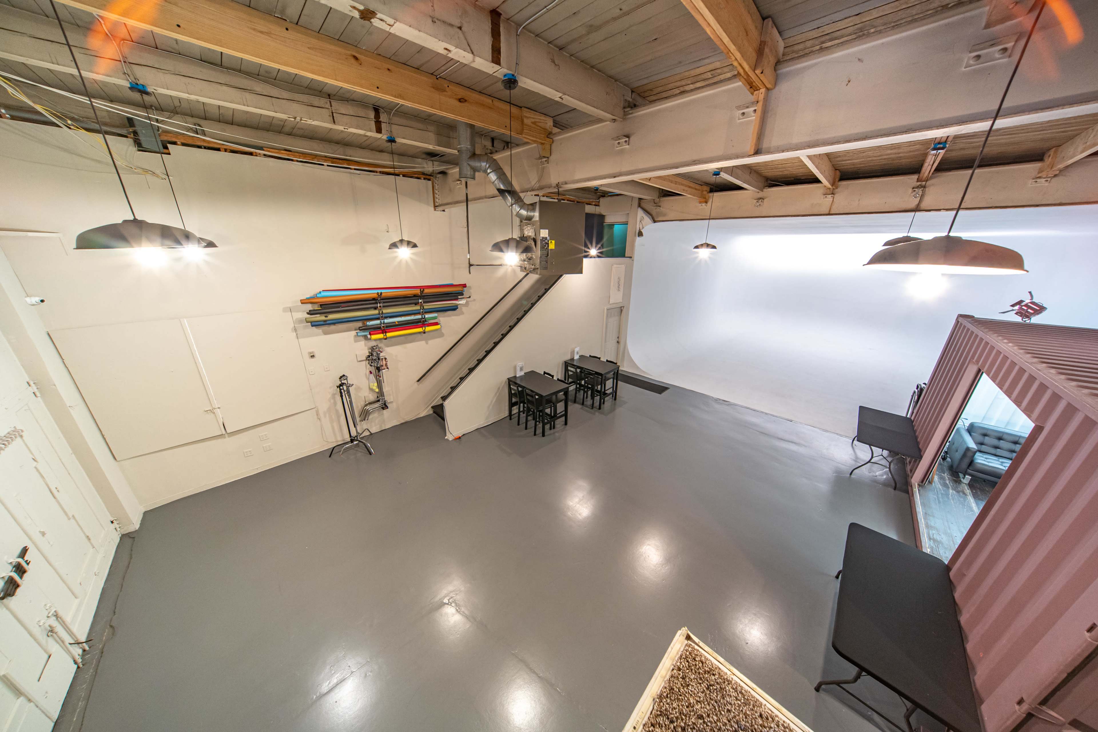 A spacious studio interior with gray flooring, overhead lights, a staircase, two tables, and a white backdrop area.
