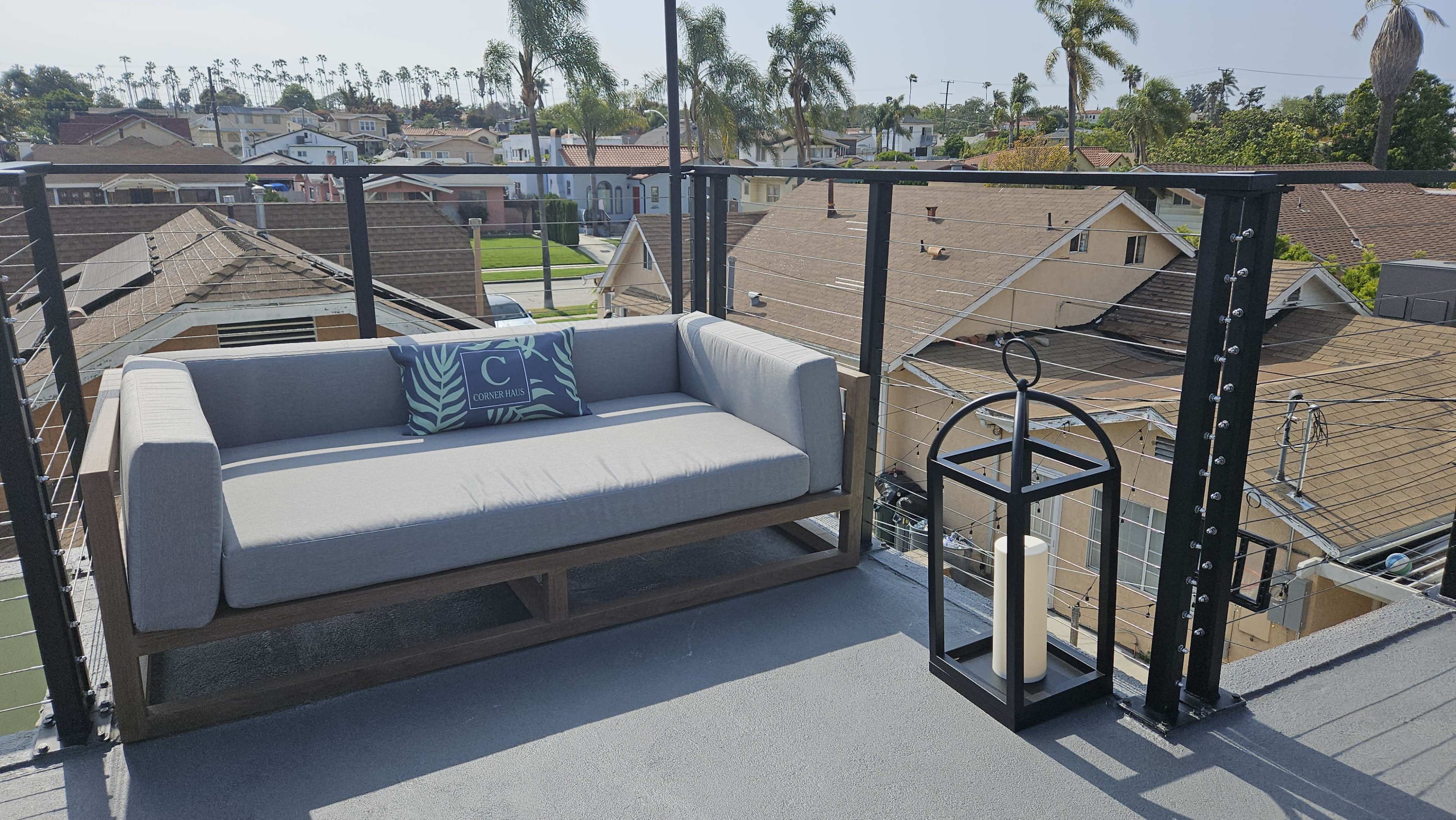 A gray outdoor sofa with a decorative pillow and a lantern stands on a balcony overlooking residential rooftops and palm trees.