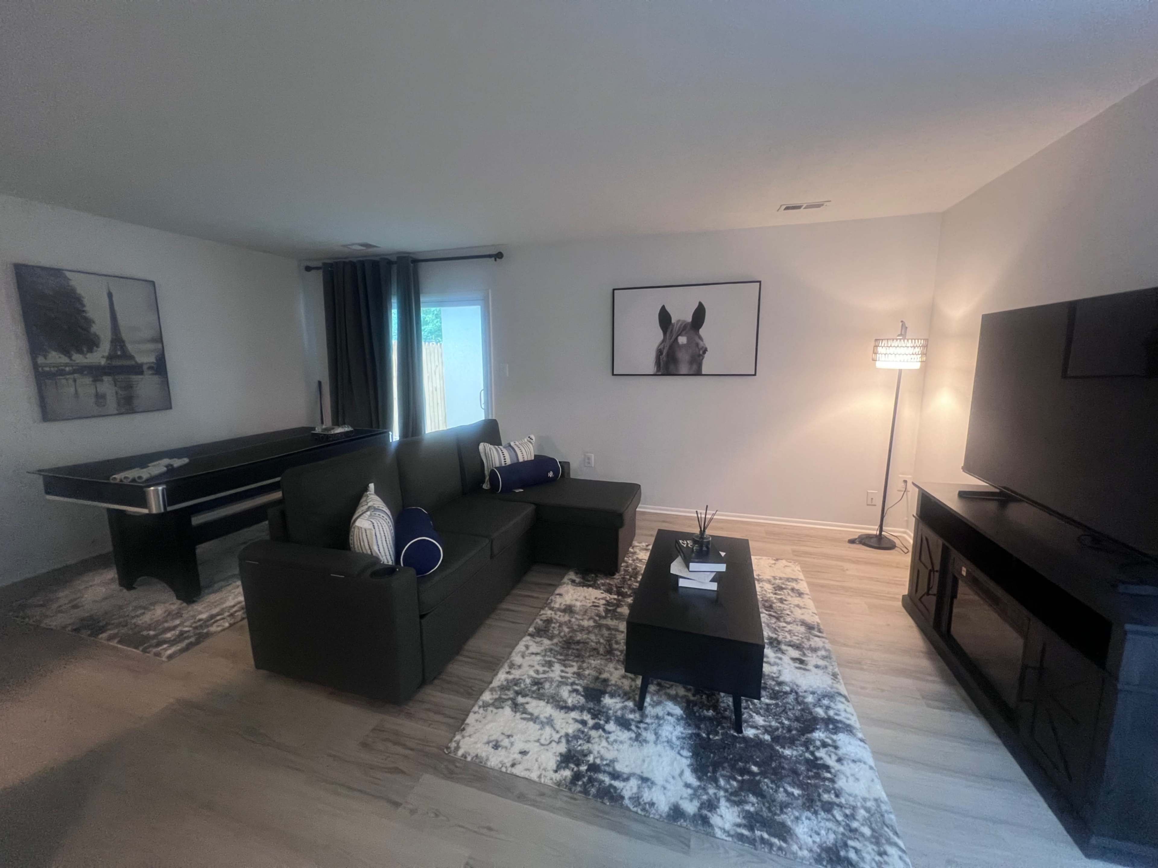 The image shows a modern living room with a gray sectional sofa, a coffee table, and a large television, alongside decorative artwork and a pool table in the background.