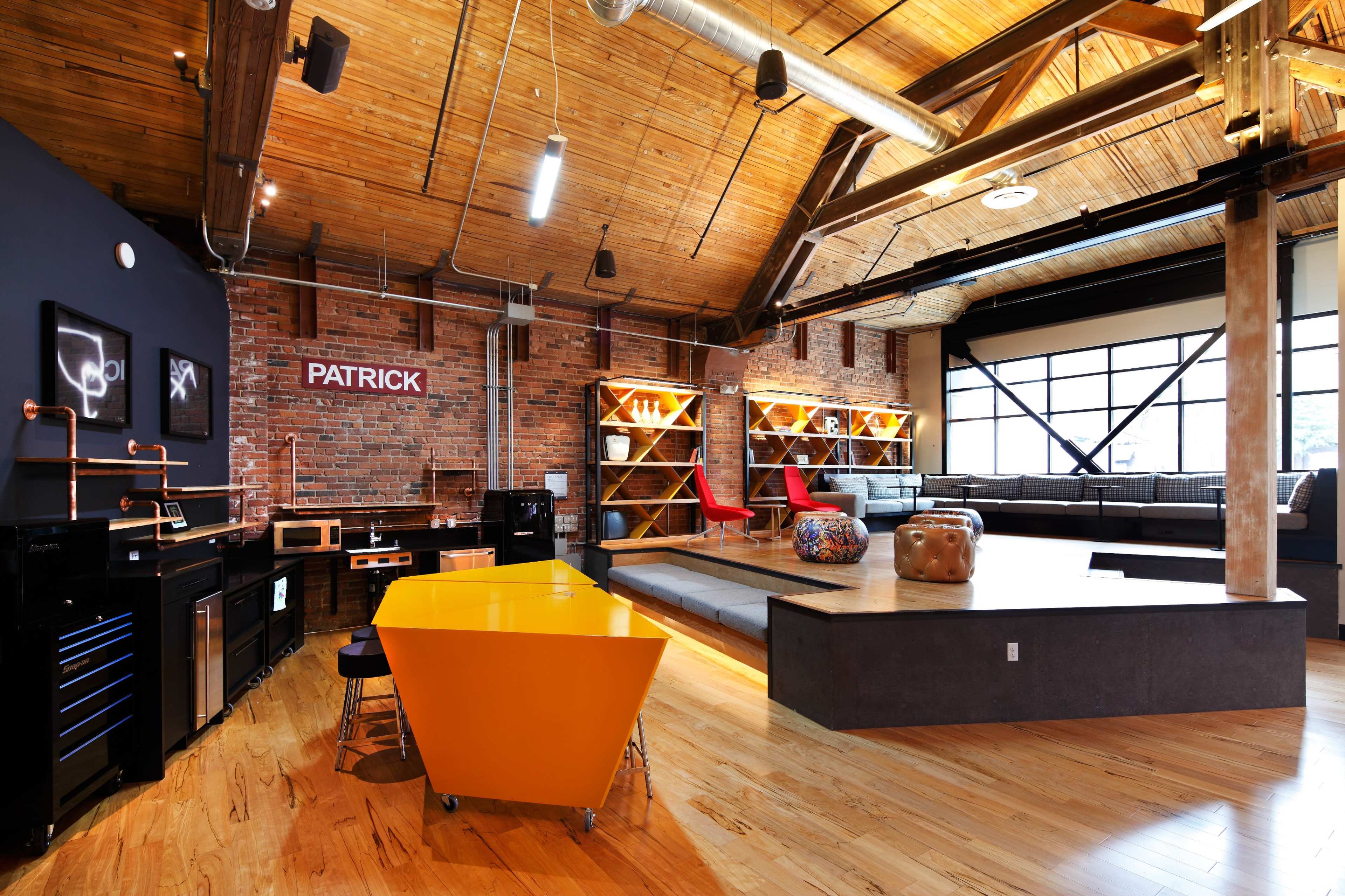 A large, open space featuring a modern kitchen area with an orange table and stools, a brick wall with the name "PATRICK," and a seating area with a sectional couch and decorative shelves.