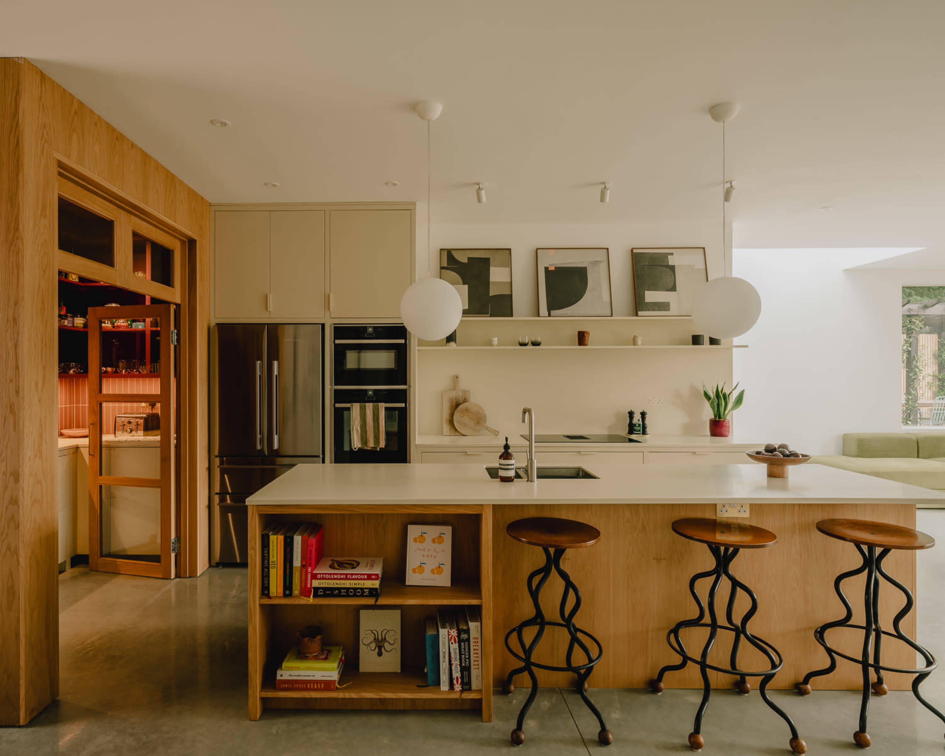 The image shows a modern kitchen featuring a central island with three wooden stools, open shelving, a refrigerator, and minimalistic decor elements.