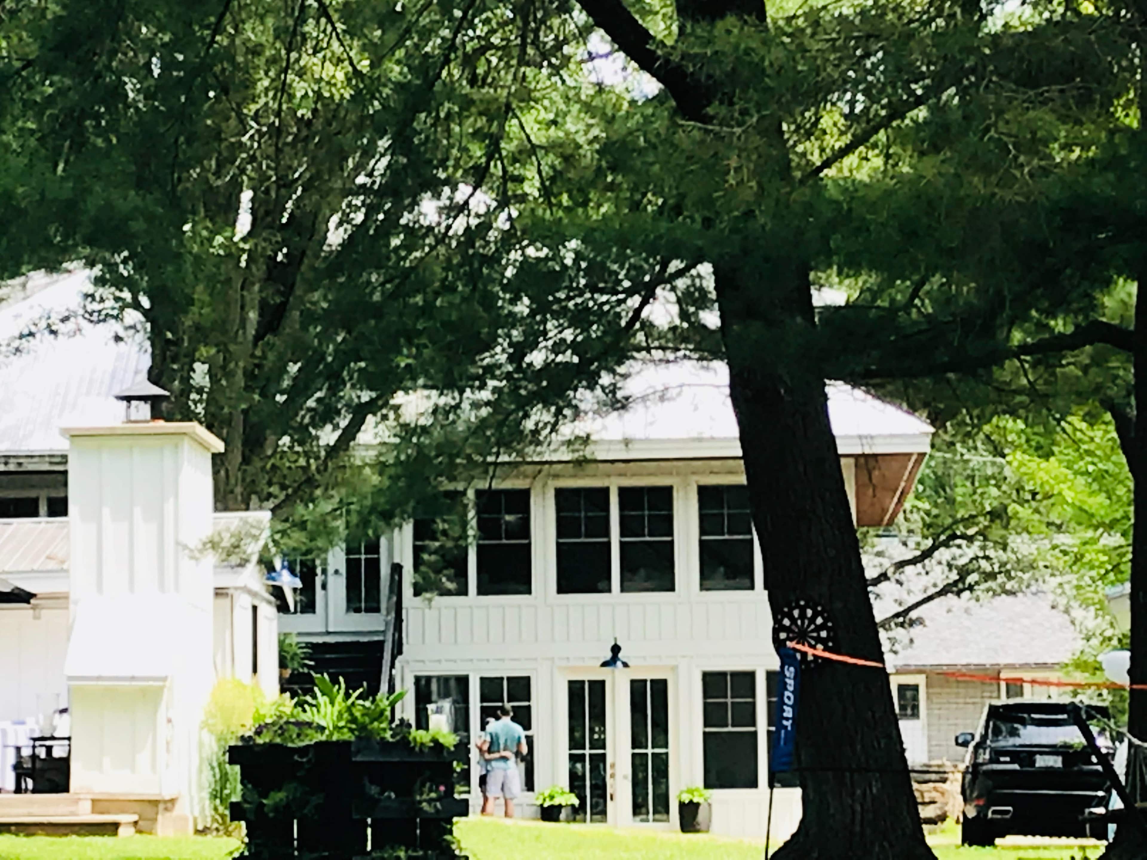 A two-story house with a white exterior and large windows is partially obscured by trees, while two people stand in front of it.