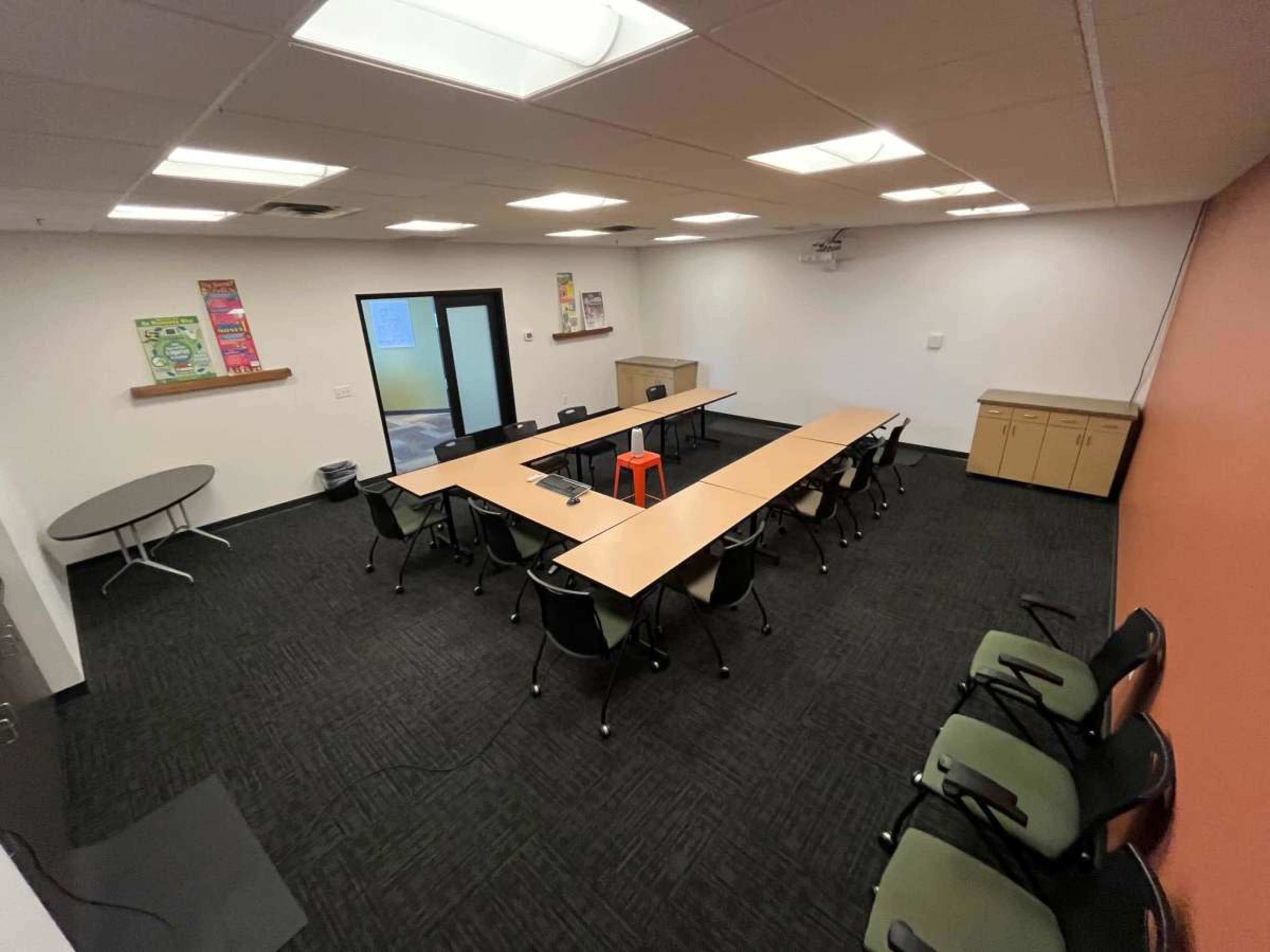 The image shows a spacious meeting room with several tables arranged in a U-shape, surrounded by chairs and a cabinet against one wall.