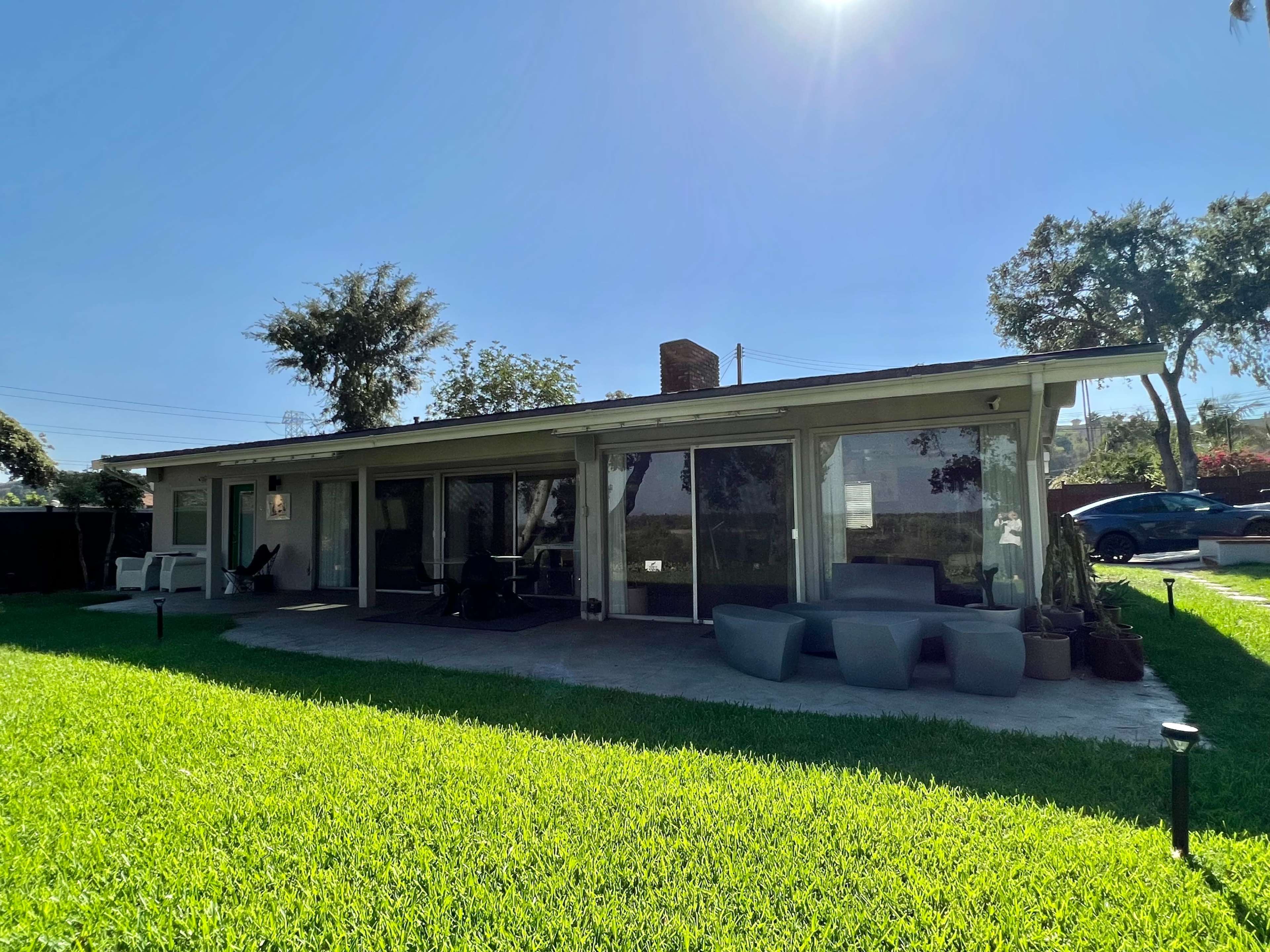 A single-story house with large windows and a patio is surrounded by a well-manicured lawn under clear blue skies.
