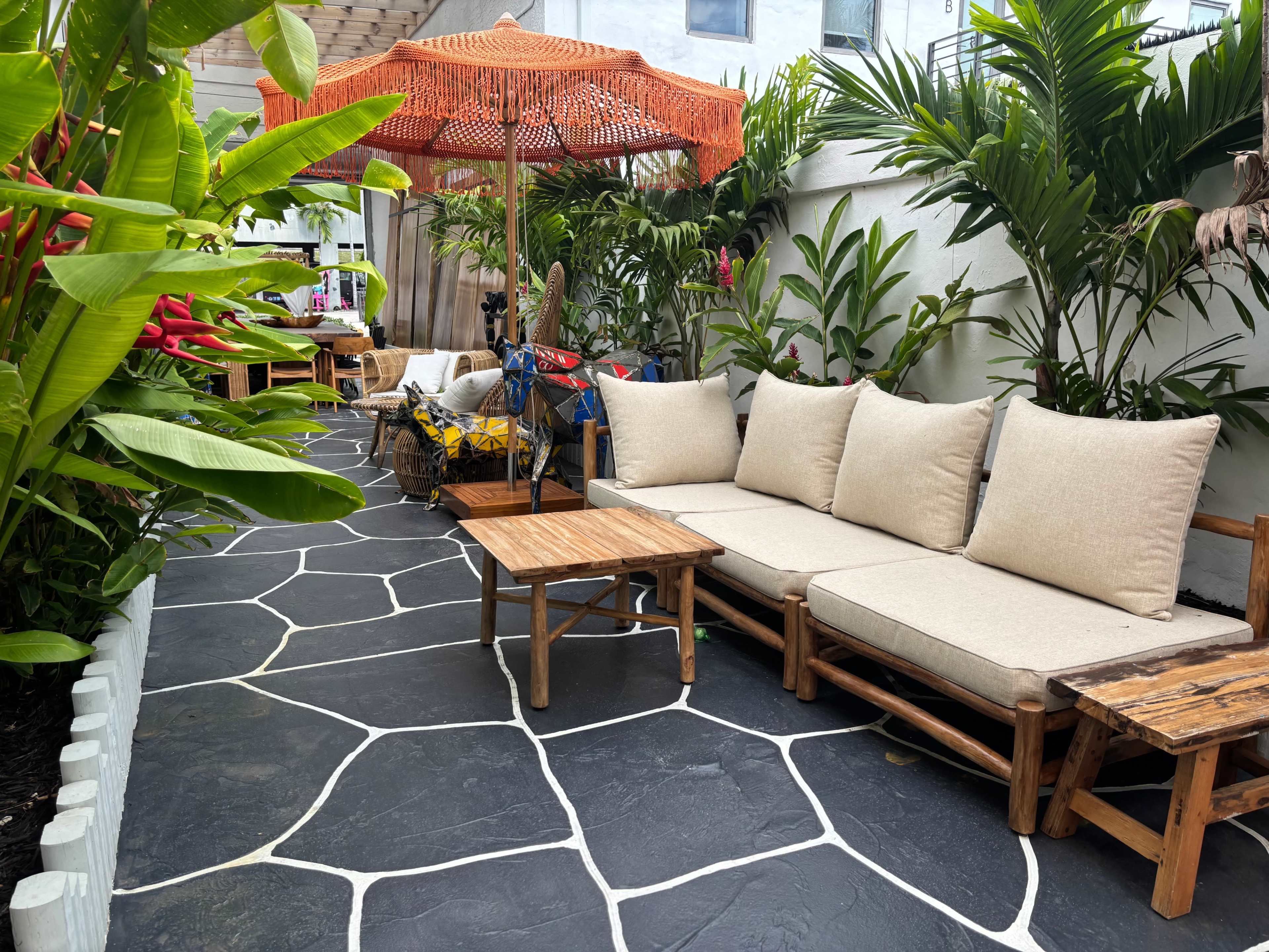 The image shows a patio area with a stone pathway, covered seating with cushions, and an umbrella, surrounded by lush green plants.