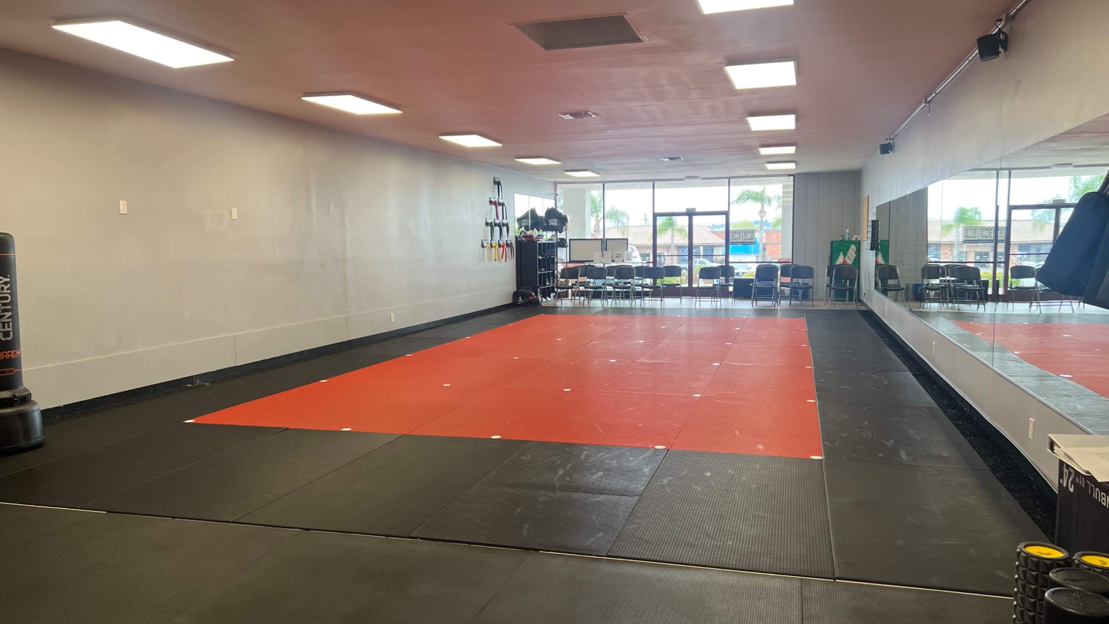 A sparsely furnished training studio with black and red mats, mirrors on one wall, and large windows on the opposite side.