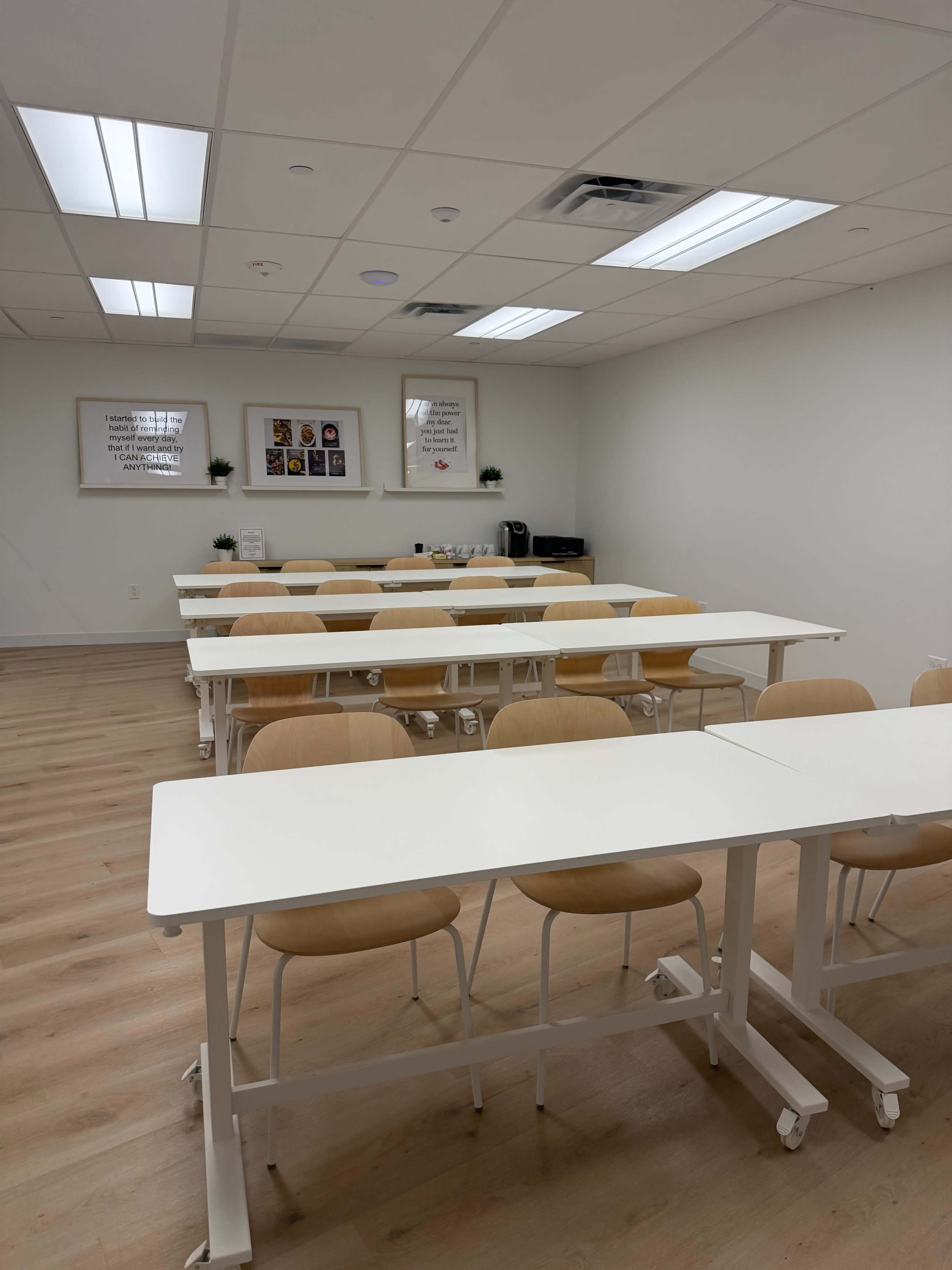 The image shows a bright, empty classroom with rows of white tables and wooden chairs, along with framed posters on the wall.