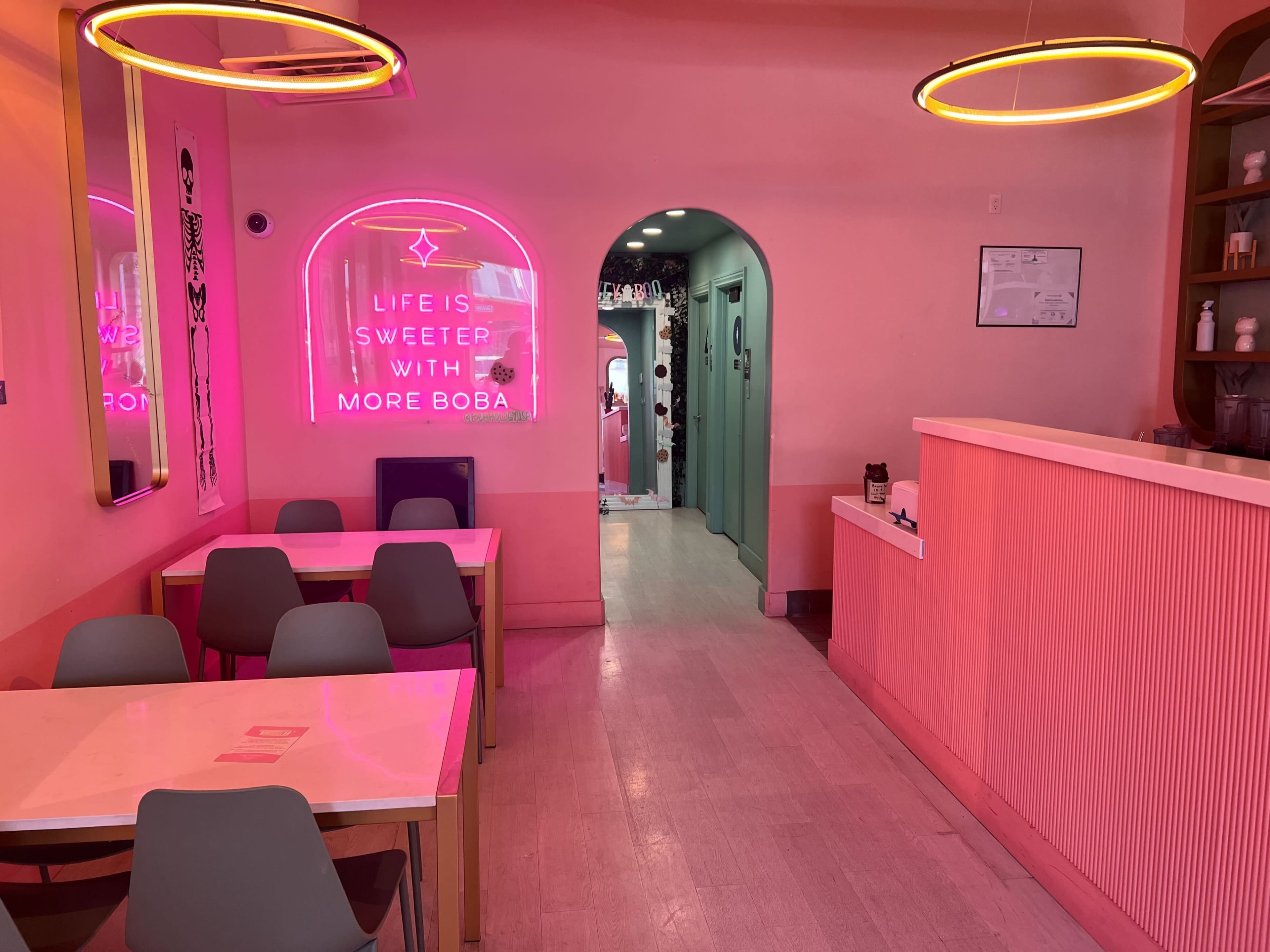 A pink-themed cafe interior with a neon sign that reads "LIFE IS SWEETER WITH MORE BOBA," featuring tables and chairs arranged around a serving area.