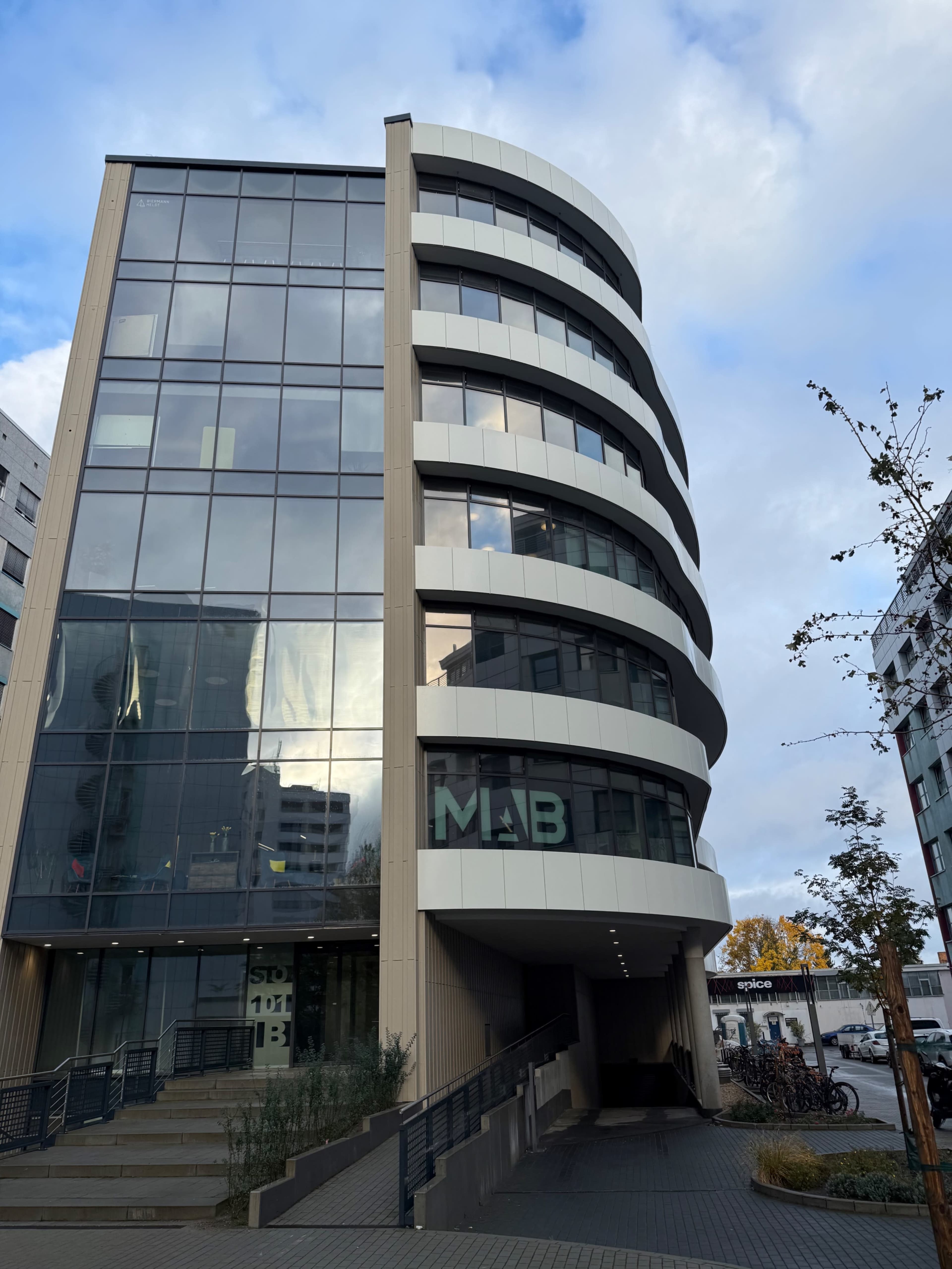 The image shows a modern, multi-story building with a curved glass facade and ramps leading to the entrance.