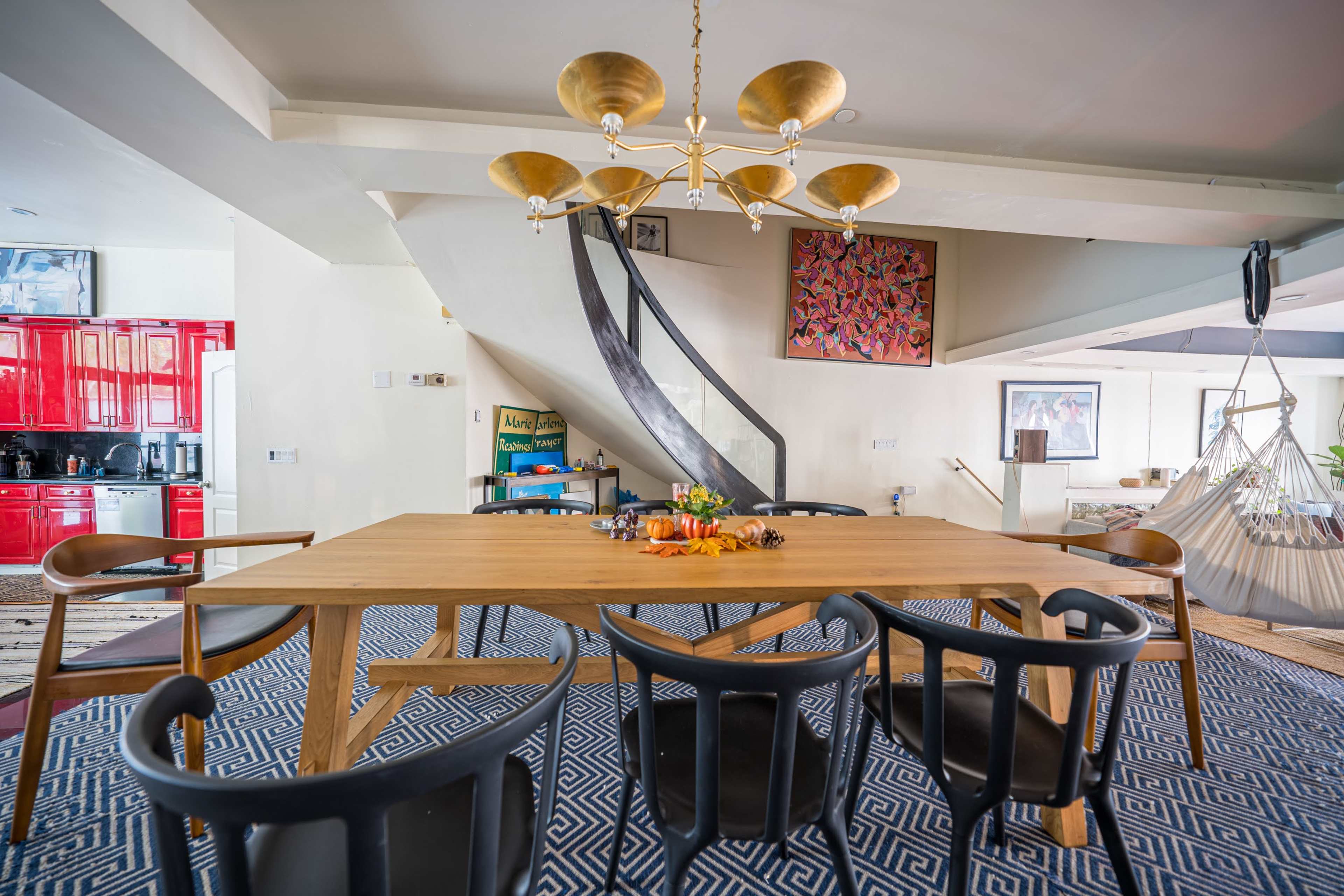 A modern dining area features a large wooden table surrounded by black chairs, with a spiral staircase in the background and kitchen visible to the left.