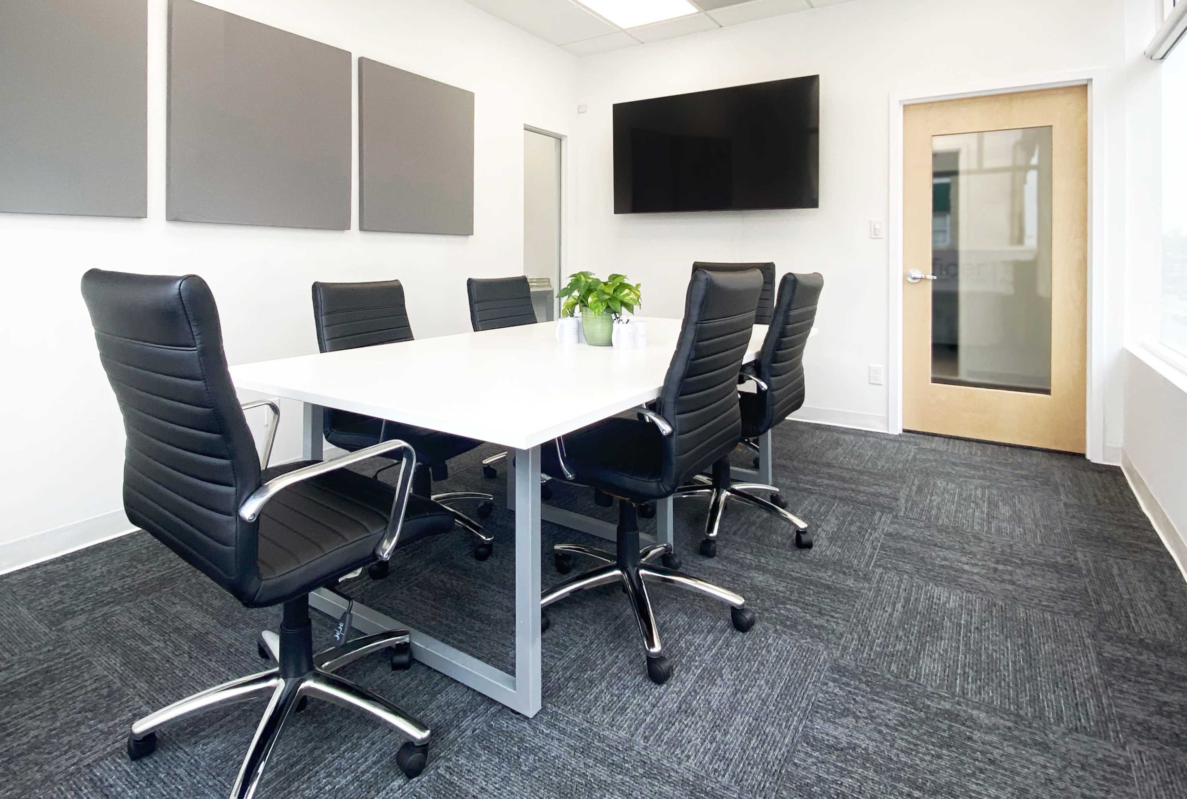 A modern conference room with a white table, black chairs, a potted plant, and a wall-mounted television.