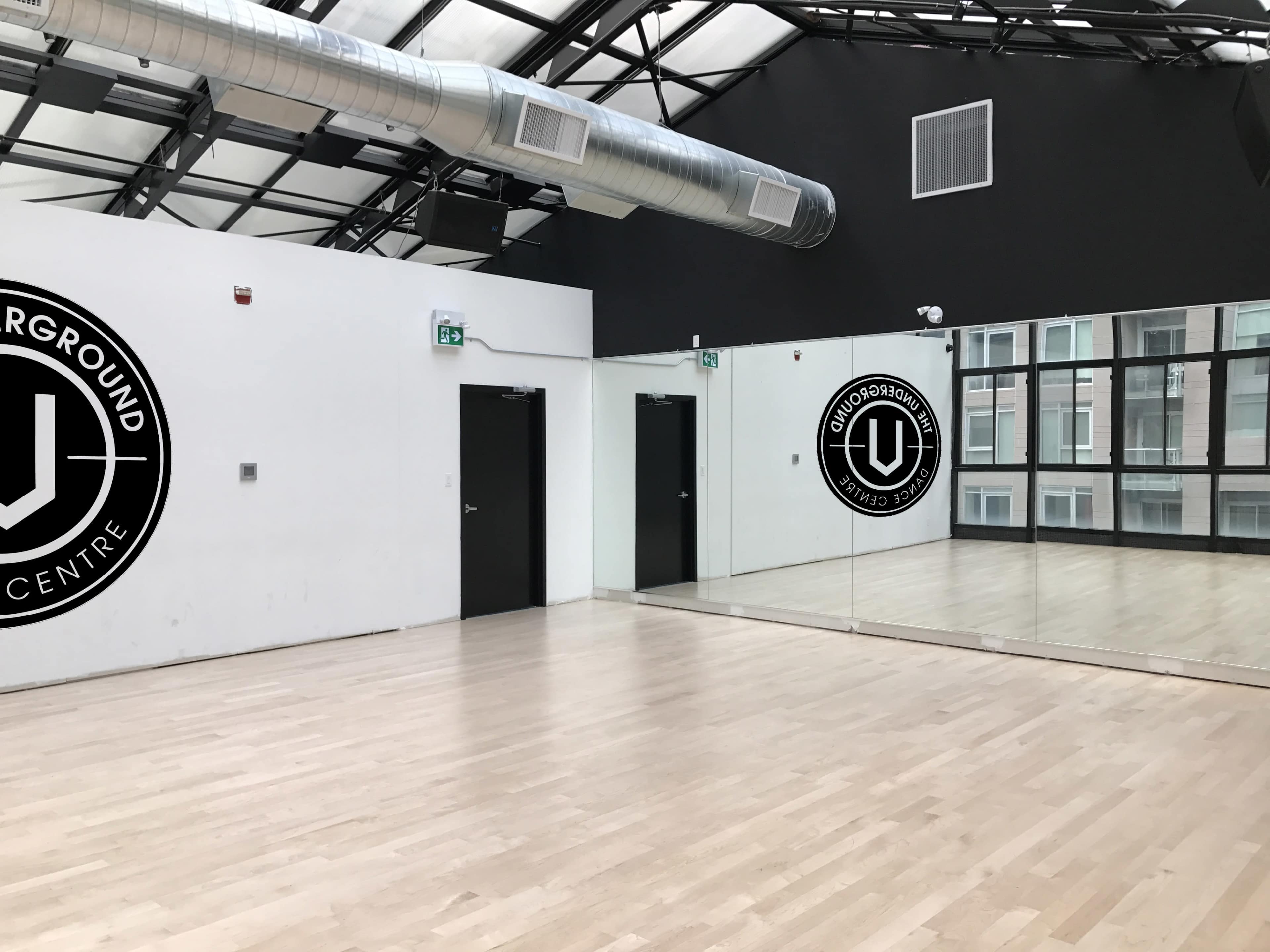 The image shows a spacious dance studio with large mirrors on one wall and a black logo on another.