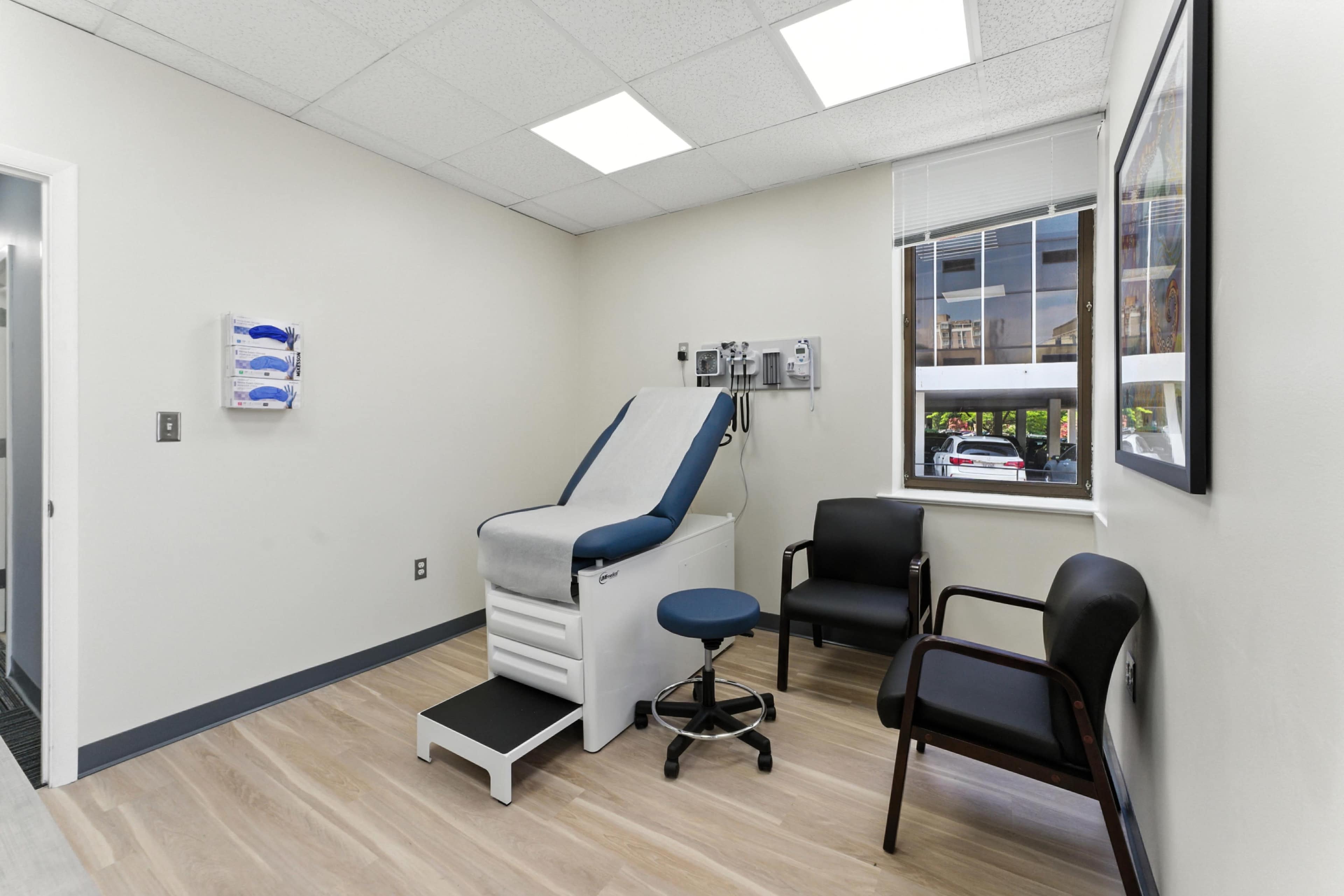 The image shows a medical examination room with an exam table, a rolling stool, and two chairs, all set against a light-colored wall with a window.