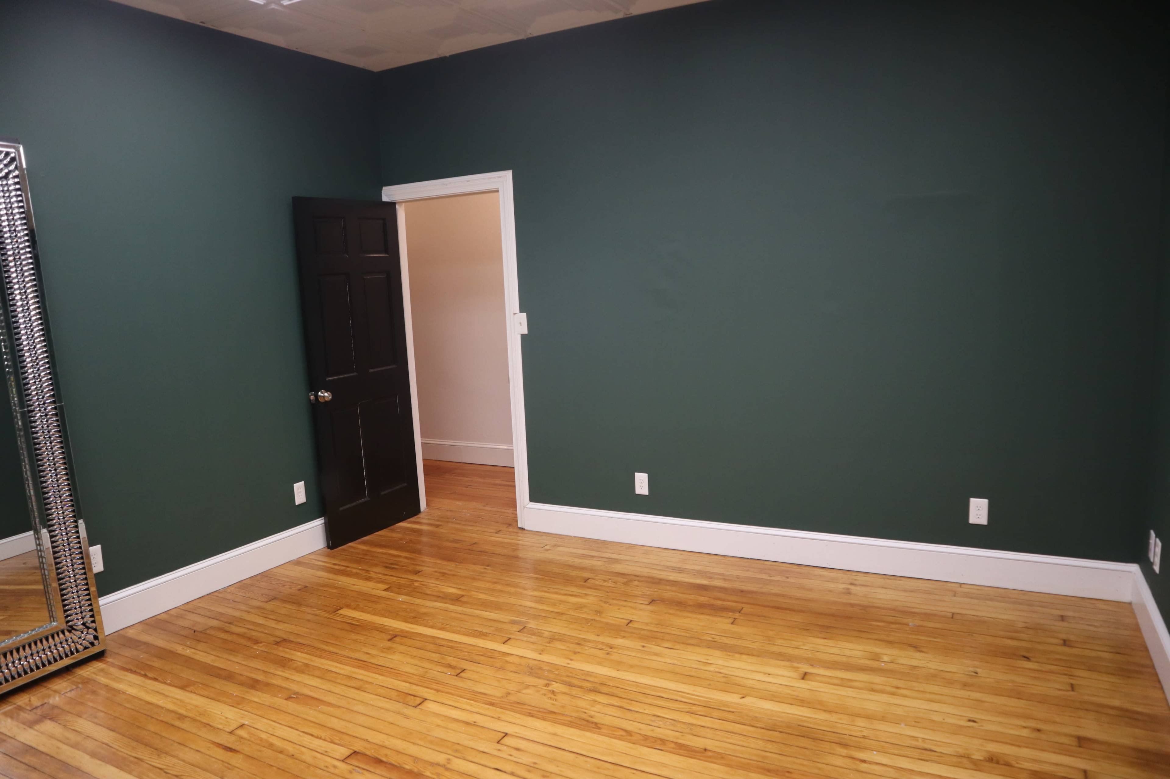 A room with dark green walls, a wooden floor, a large mirror on one side, and a black door leading to another space.