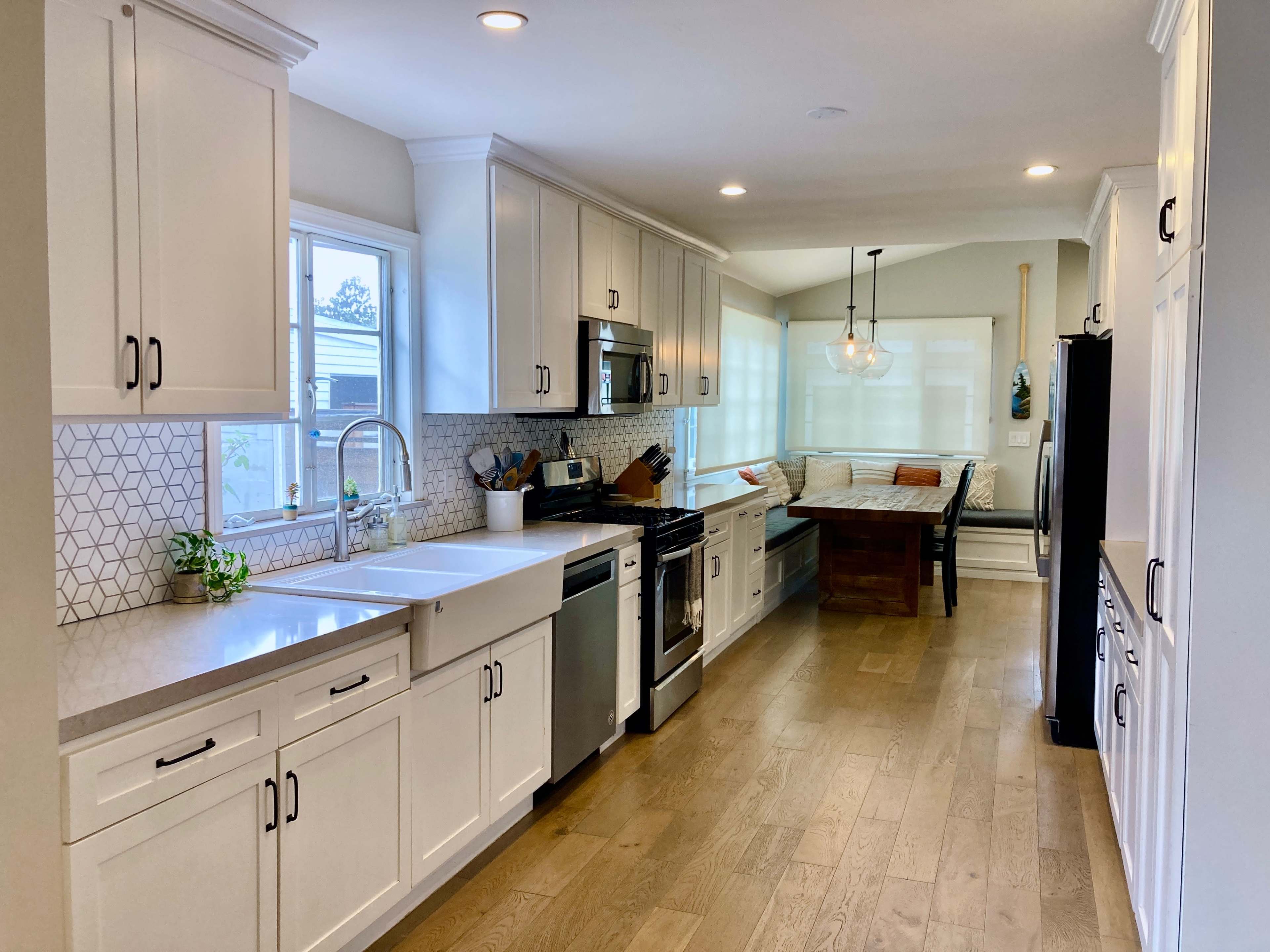 The image shows a modern kitchen with white cabinetry, a large sink, a black stove, a dining table, and natural light coming through the windows.