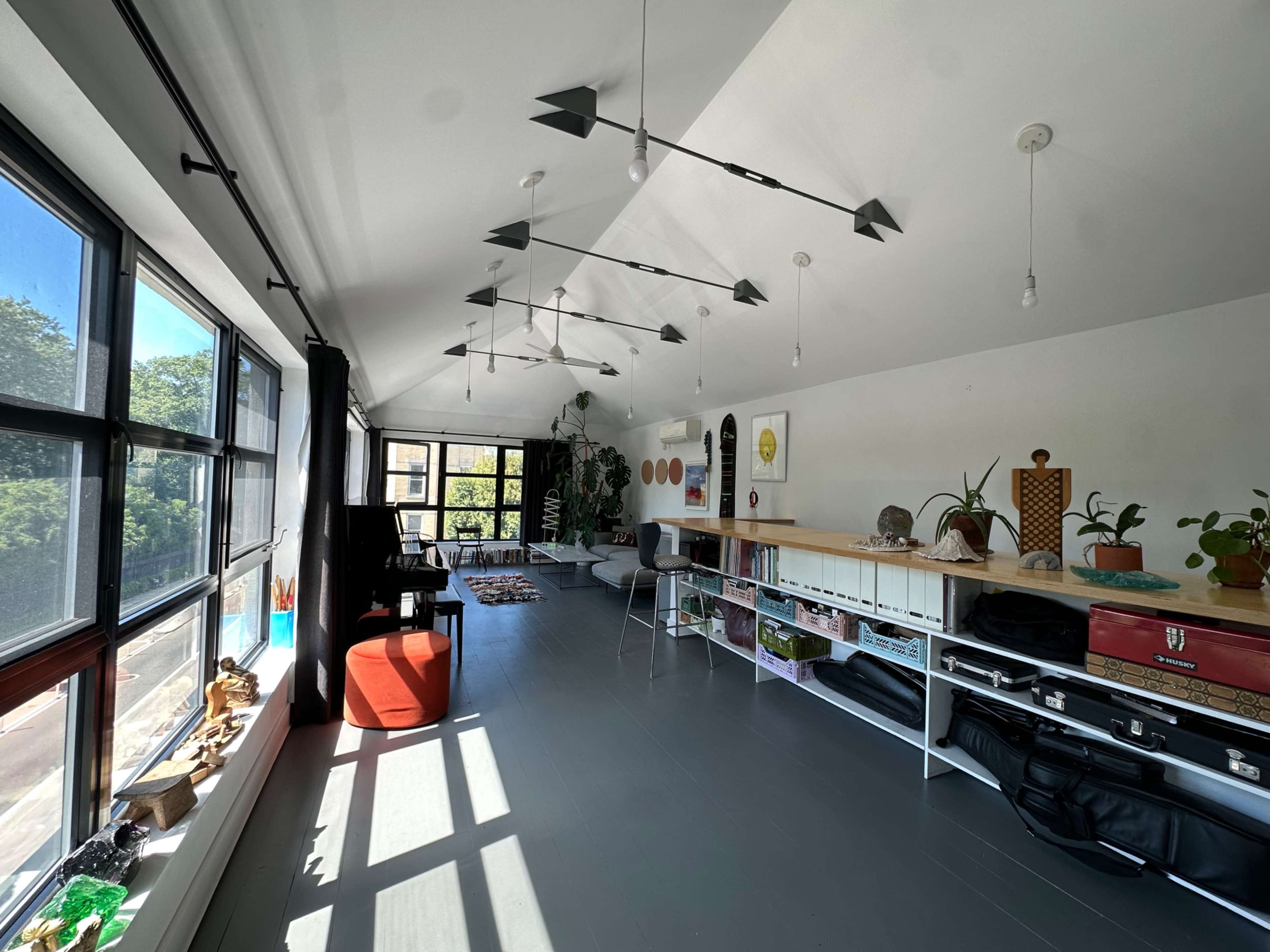 The image shows a bright, modern open space with large windows, wooden flooring, and various plants, artwork, and furniture arranged along the walls.