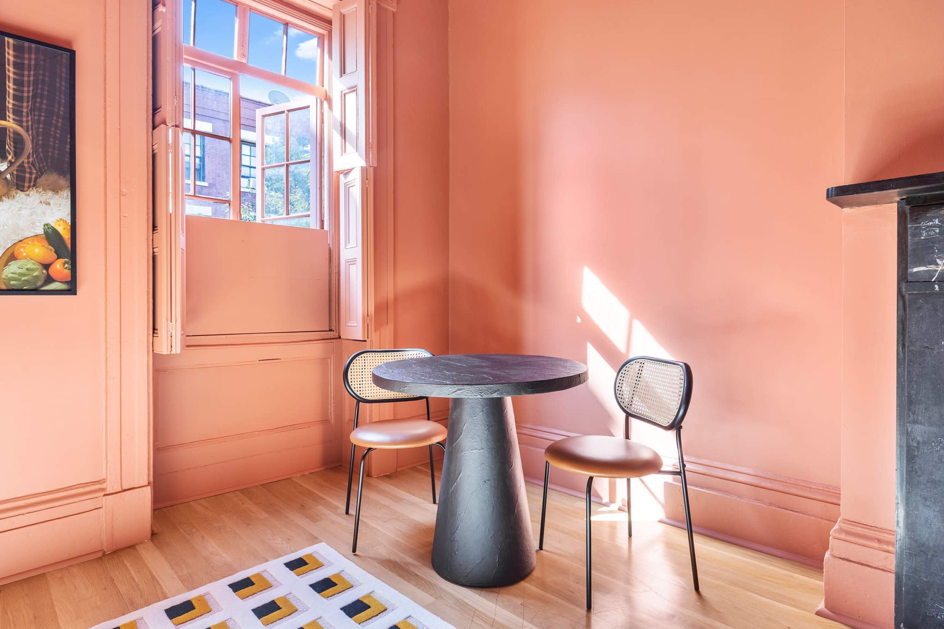 A small dining area features a round black table and two chairs against a light pink wall with an open window.