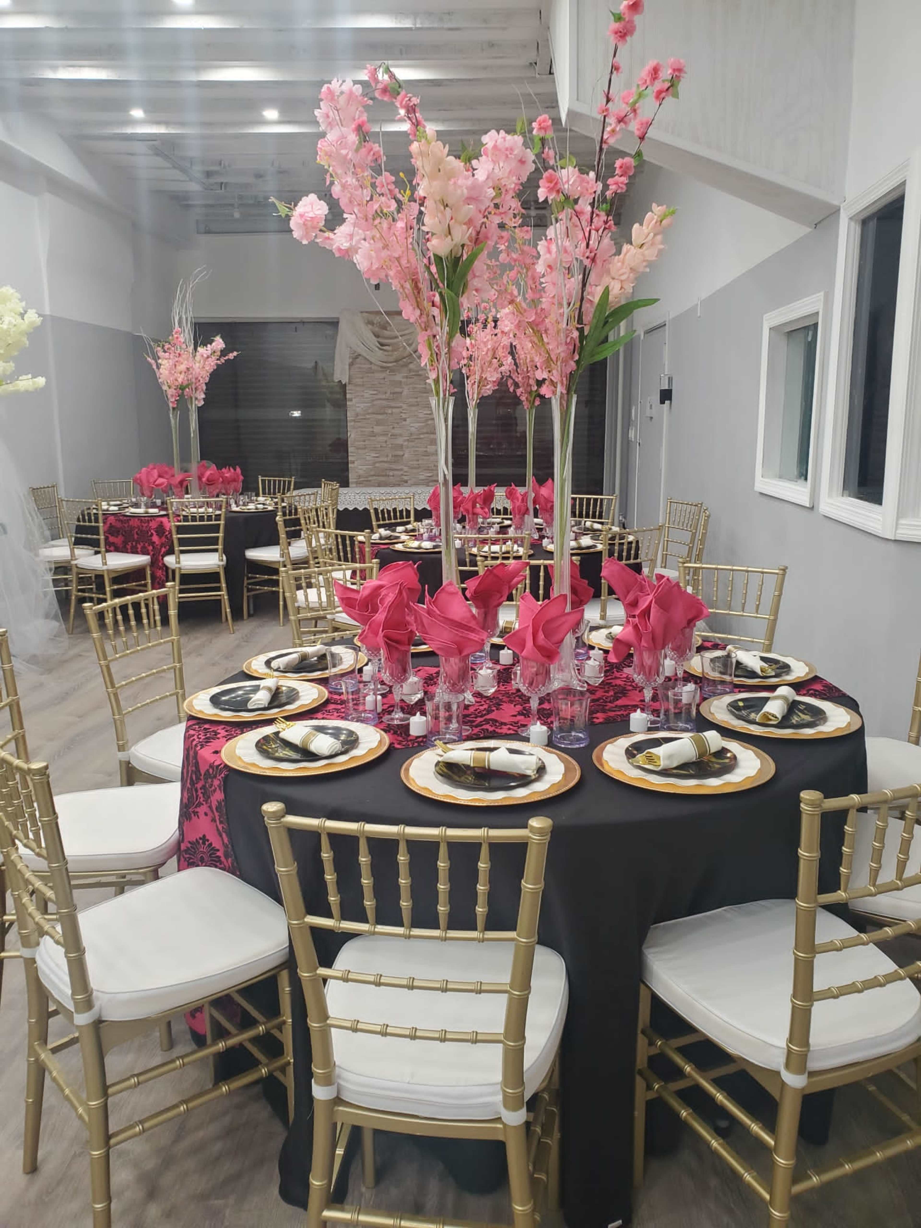 A banquet hall is set up with elegantly arranged tables featuring black and gold table settings, pink floral centerpieces, and white chairs.