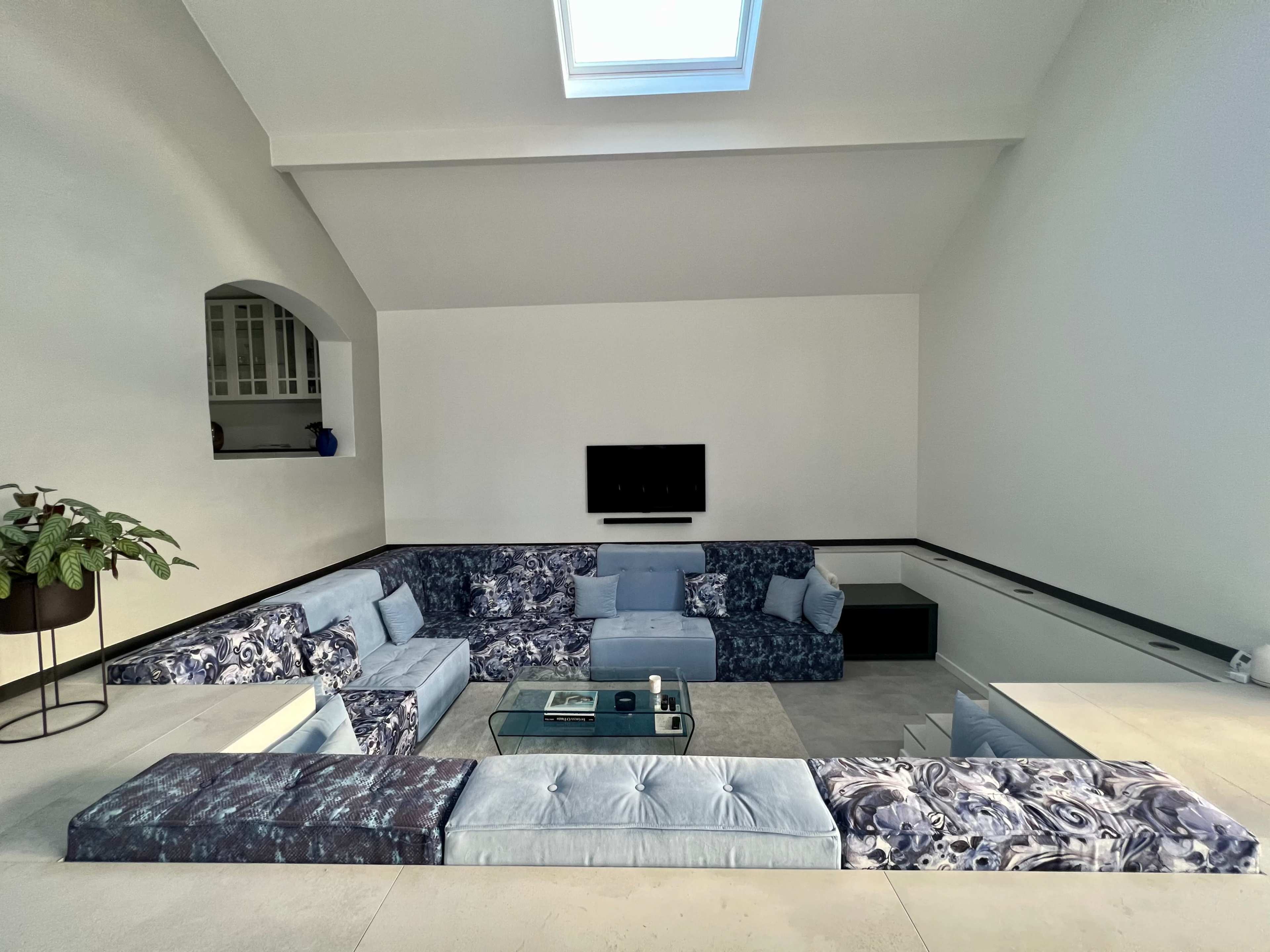 A modern living room featuring a sectional sofa in blue tones, a glass coffee table, and large windows providing natural light.