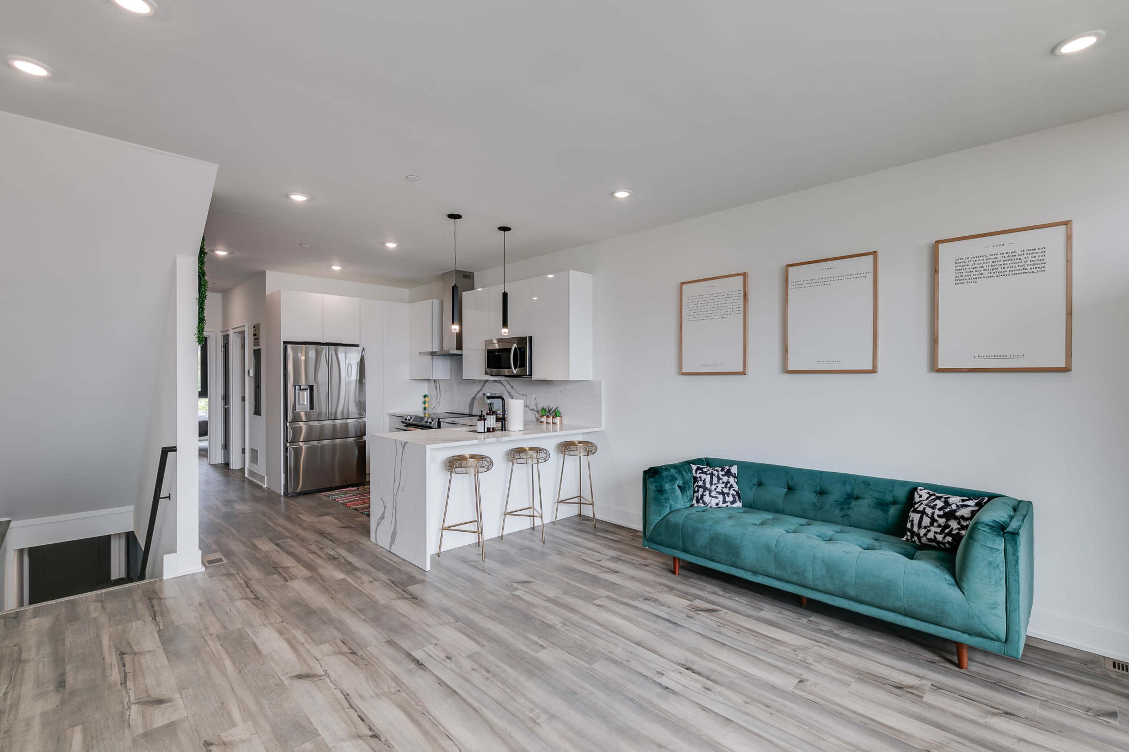 A modern, open-concept kitchen and living space featuring a turquoise sofa, a kitchen island with bar stools, and sleek stainless steel appliances.