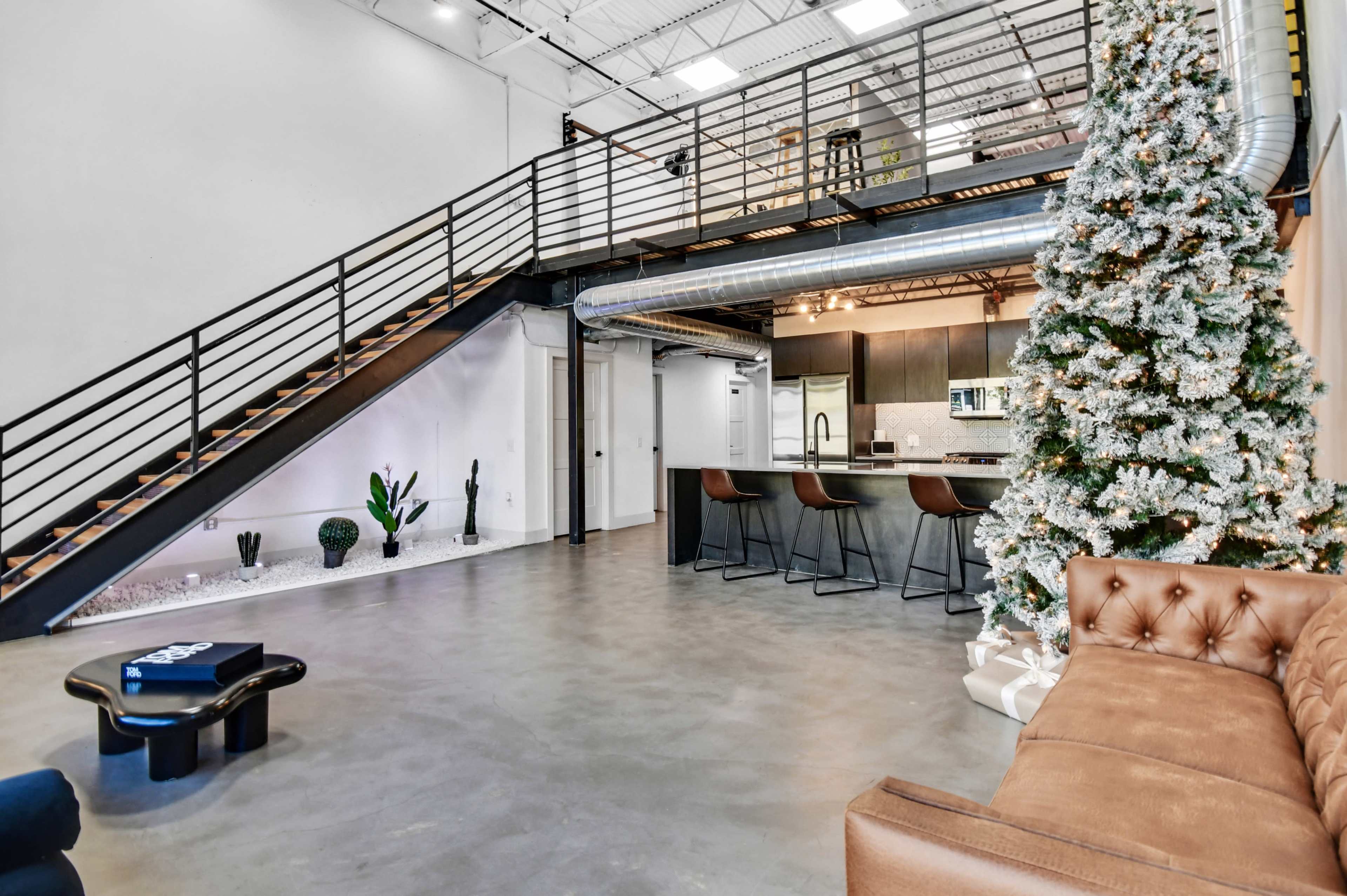 The image shows a modern industrial-style interior with a staircase, a cozy seating area, a kitchen space, and a decorated Christmas tree.