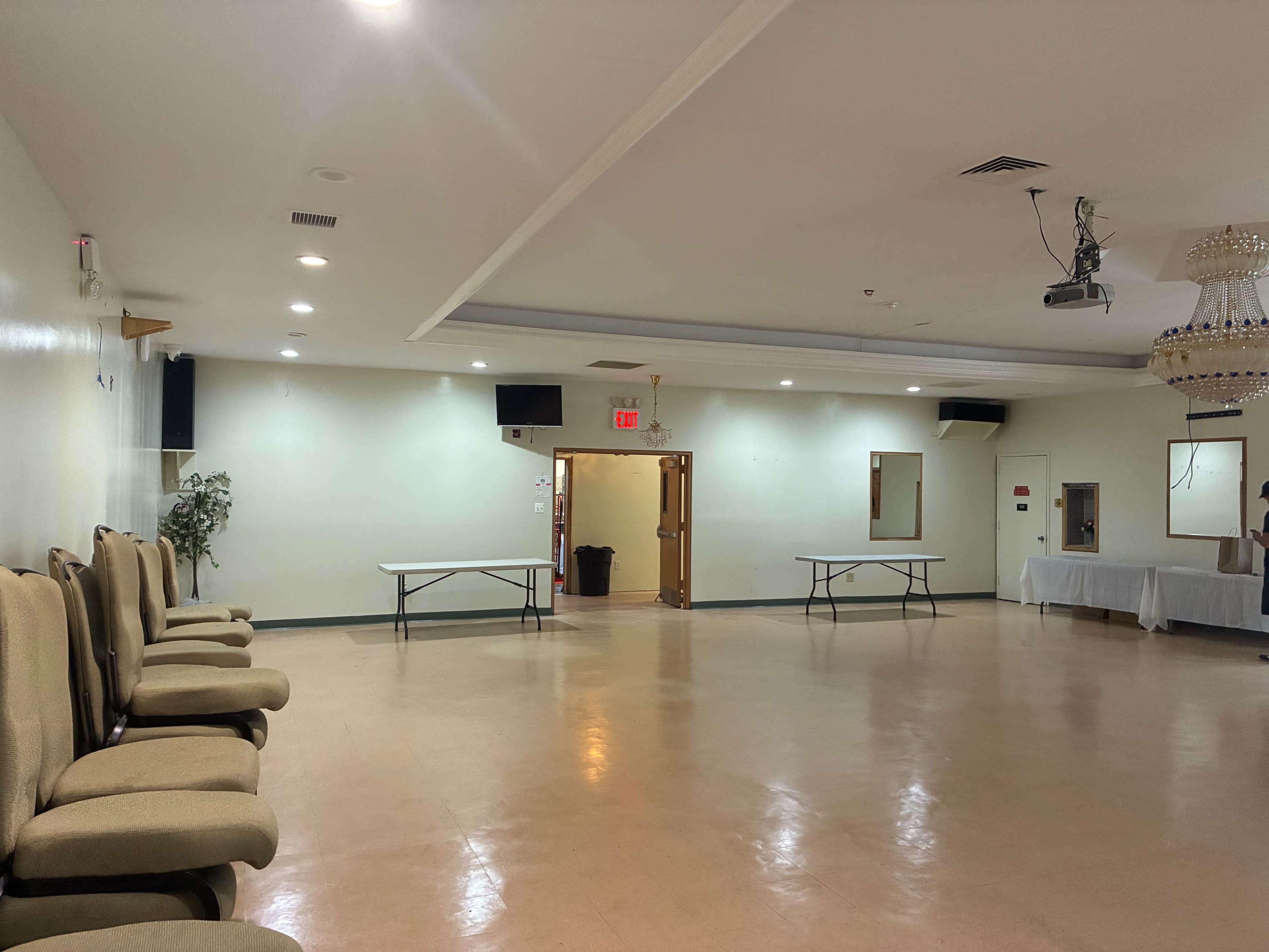 The image shows a spacious, empty hall with beige walls, a chandelier, and several folding tables arranged in the center.