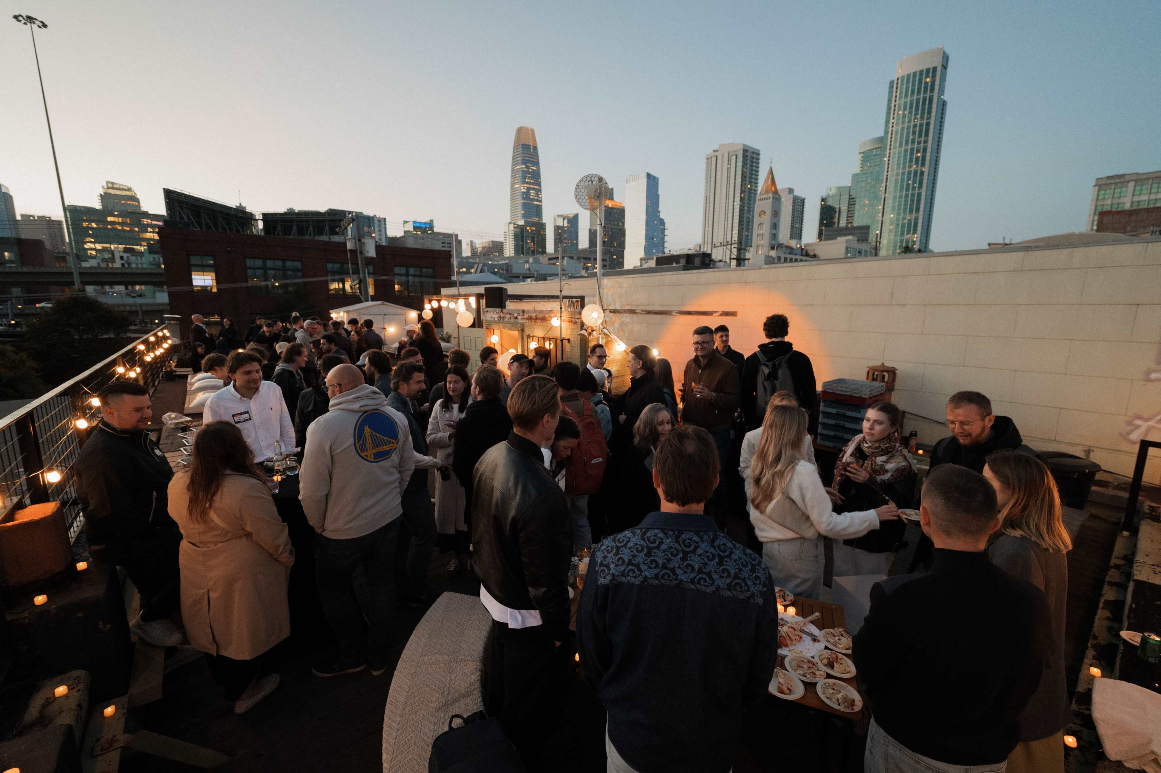 A crowd gathers on a rooftop with city skyline views, enjoying food and drinks during an evening event.