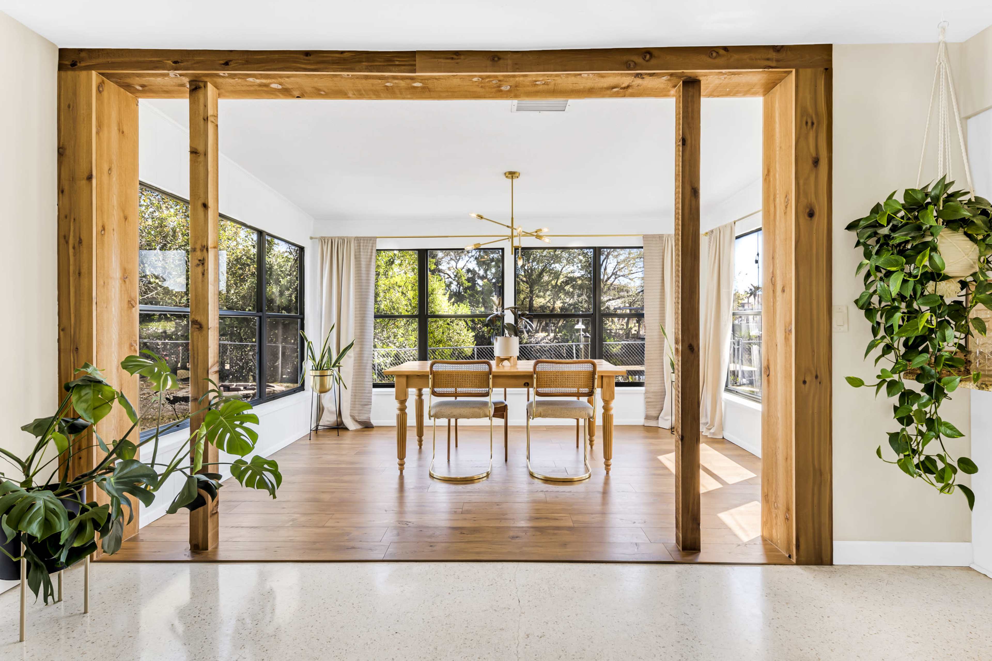A spacious dining area features a wooden table surrounded by chairs, framed by large windows and a view of greenery outside.