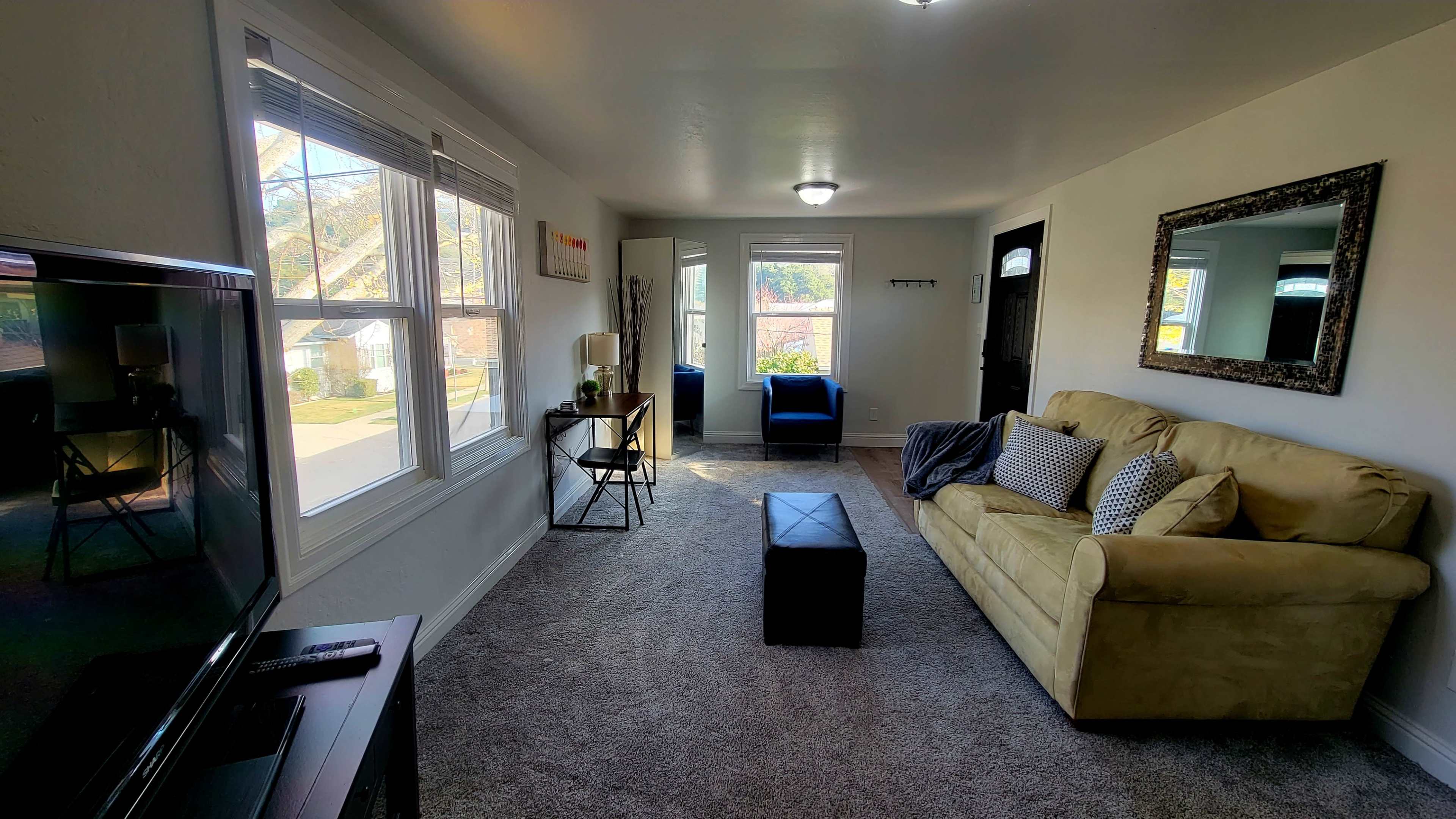 The room features a beige sofa, a dark ottoman, a television, and large windows providing natural light, with a mirror and a chair visible in the background.