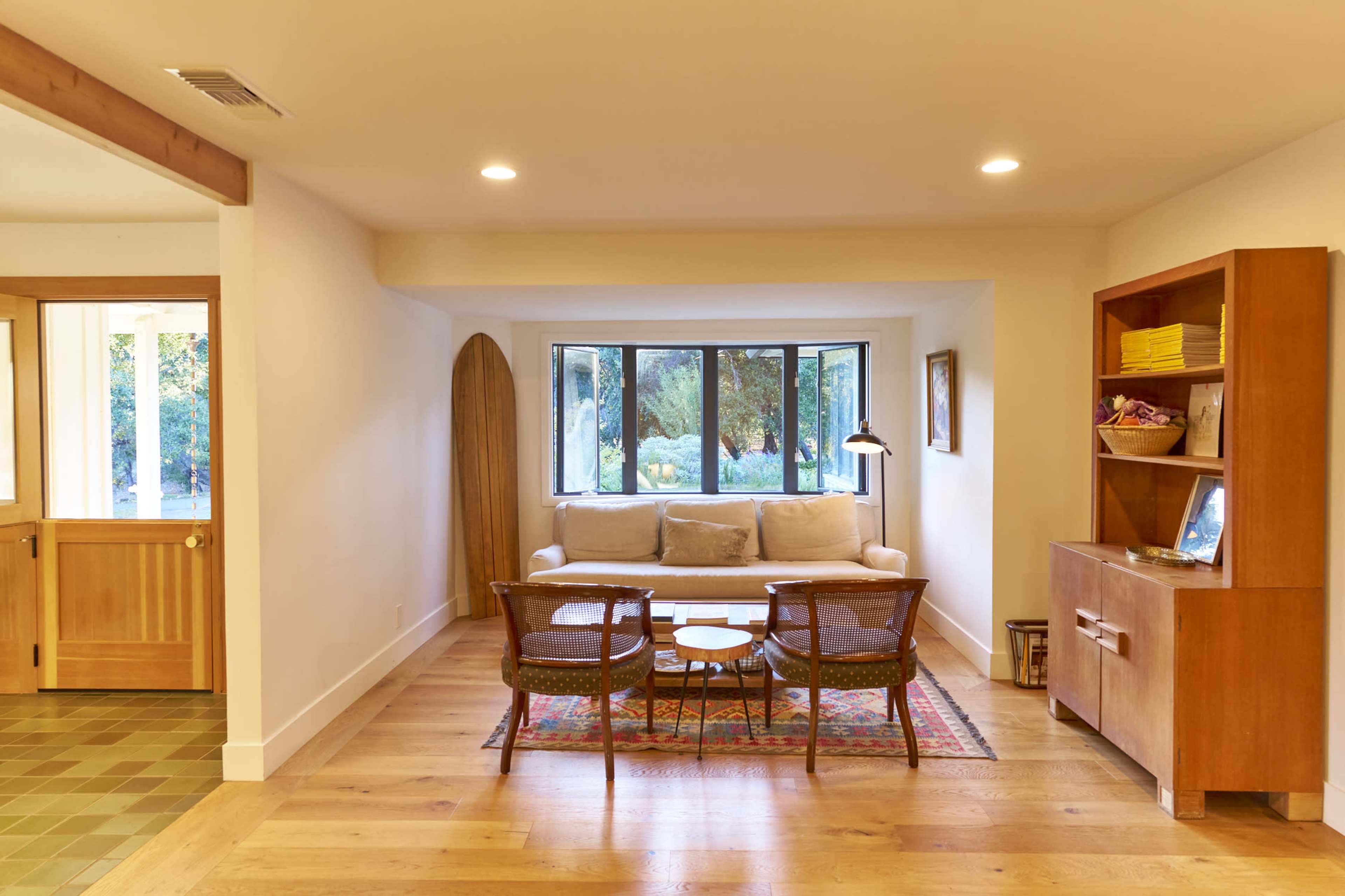 A cozy living room features a light-colored sofa and a coffee table, with large windows overlooking a garden, and wooden furniture lining the walls.