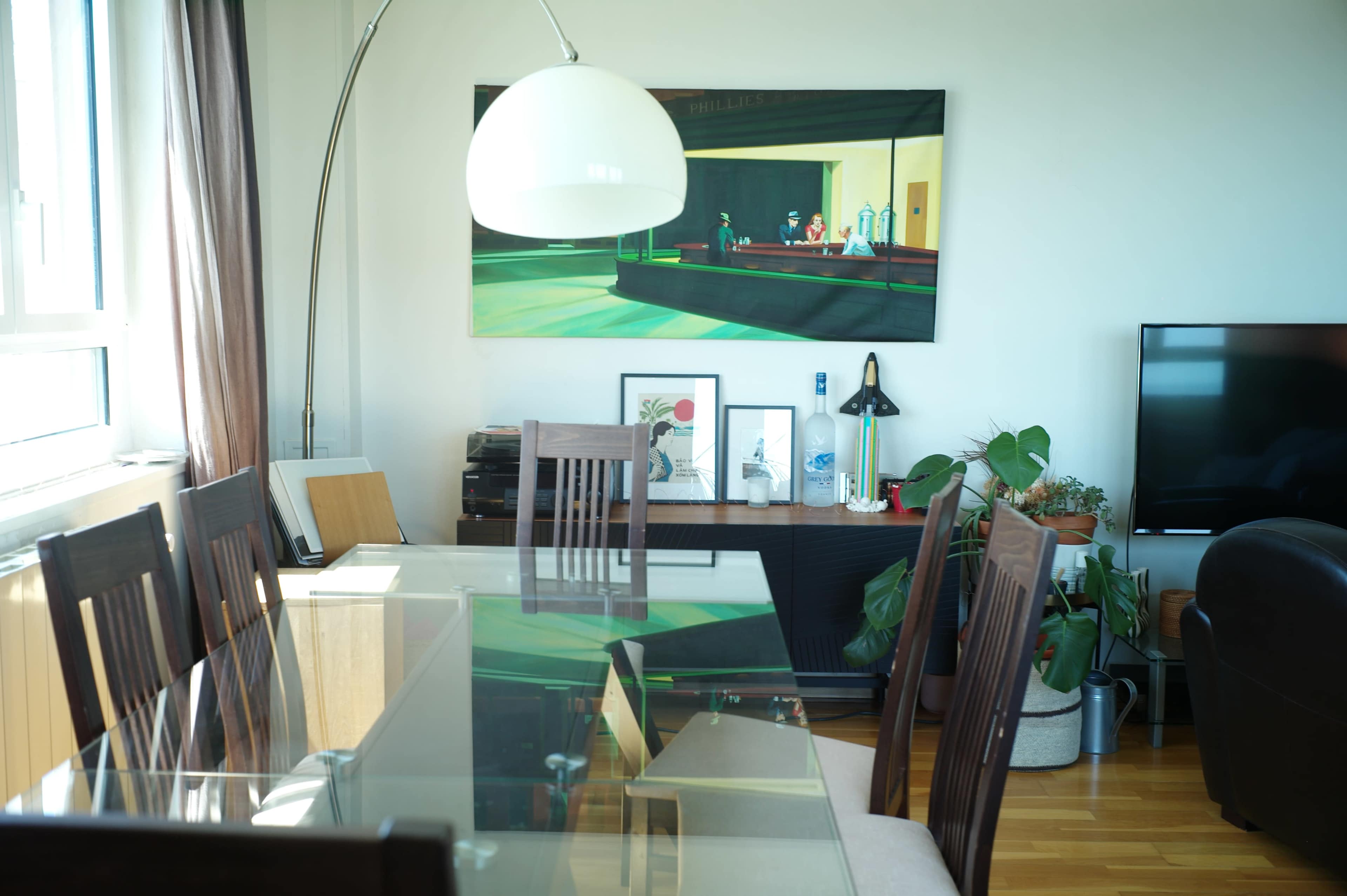 The image shows a modern dining area featuring a glass dining table, wooden chairs, a large painting on the wall, and various decorative elements, including plants and a lamp.