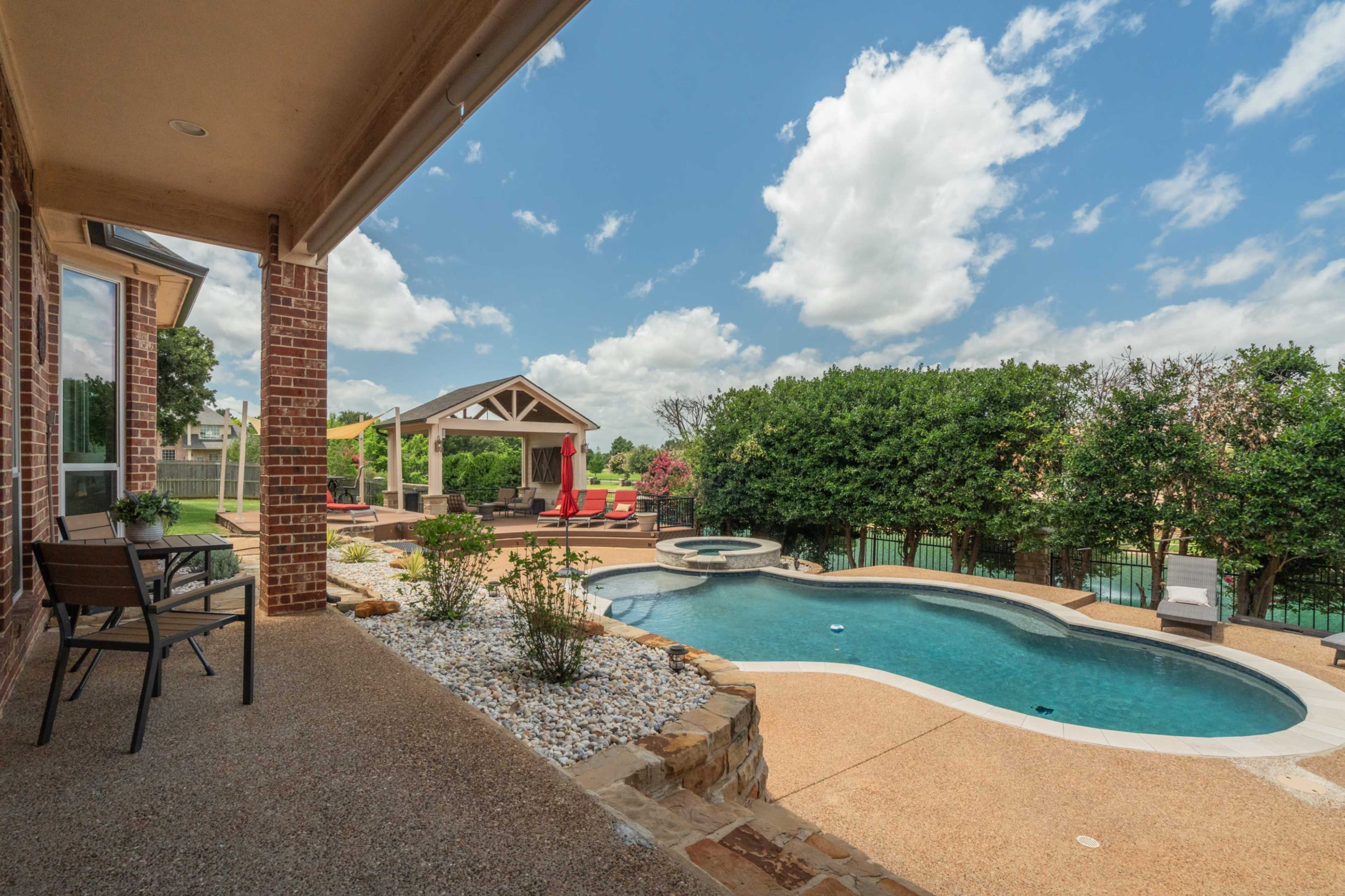 The image shows a well-maintained backyard with a swimming pool, a covered outdoor seating area, and landscaped greenery.