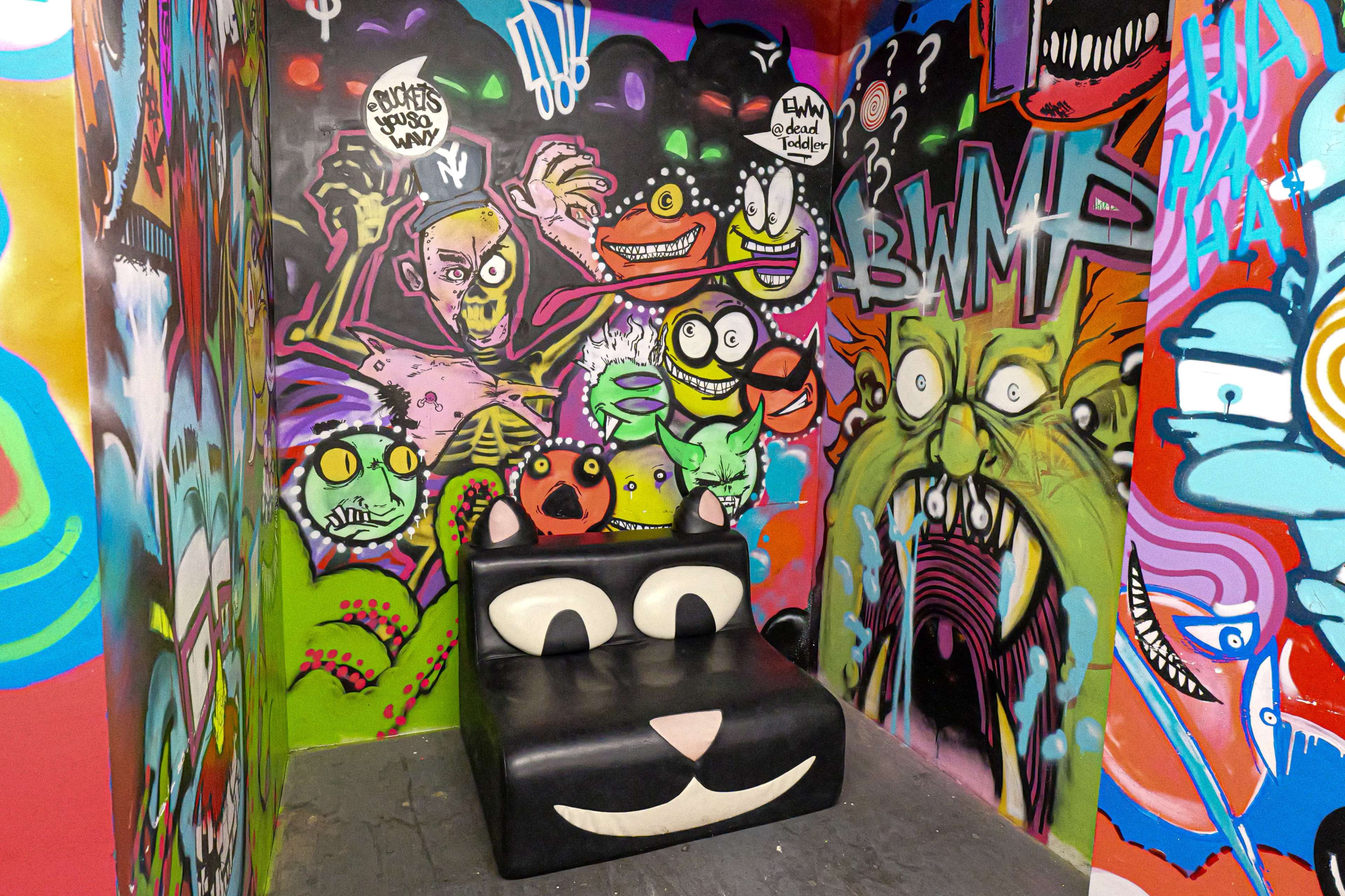 The image features a colorful, graffiti-covered room with a black cat-shaped couch in the center.