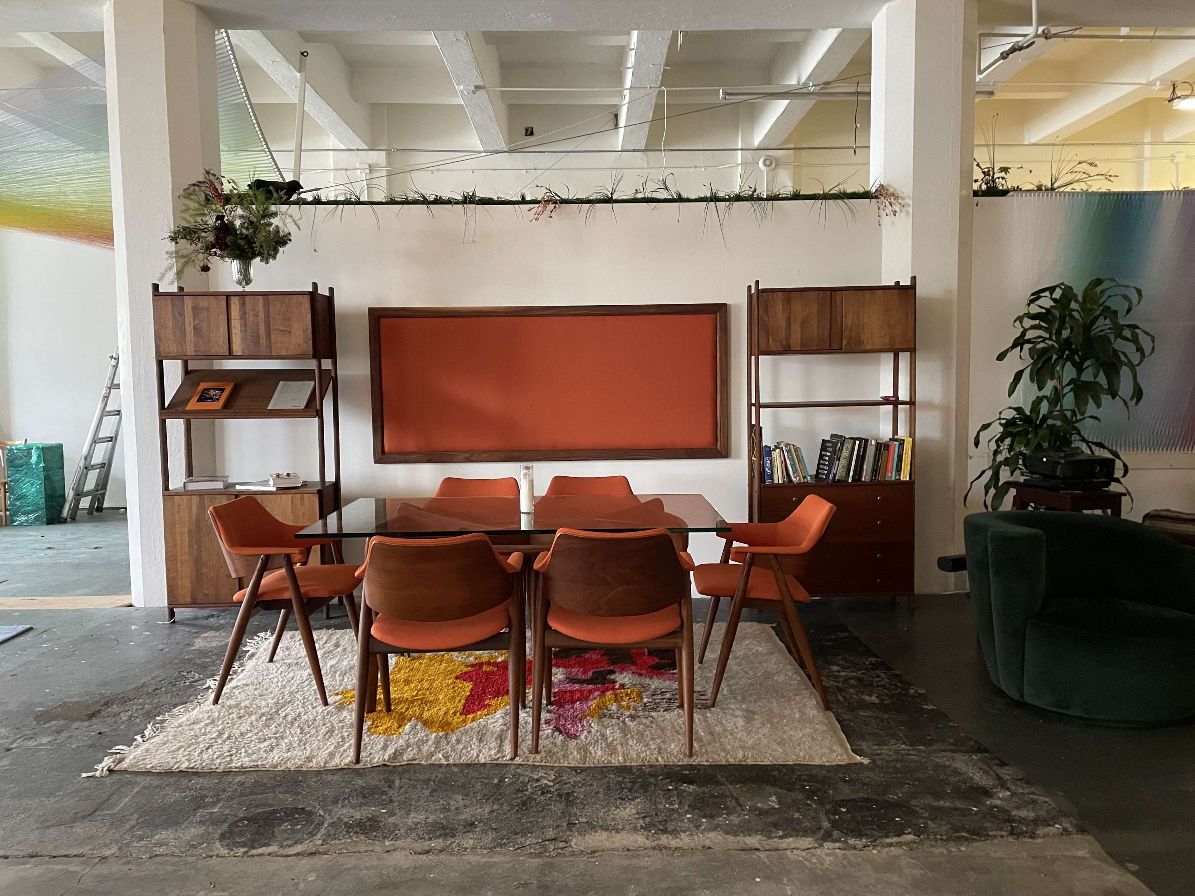 A dining area features a wooden table surrounded by six chairs on a patterned rug, with a bookshelf and a wall-mounted display in the background.