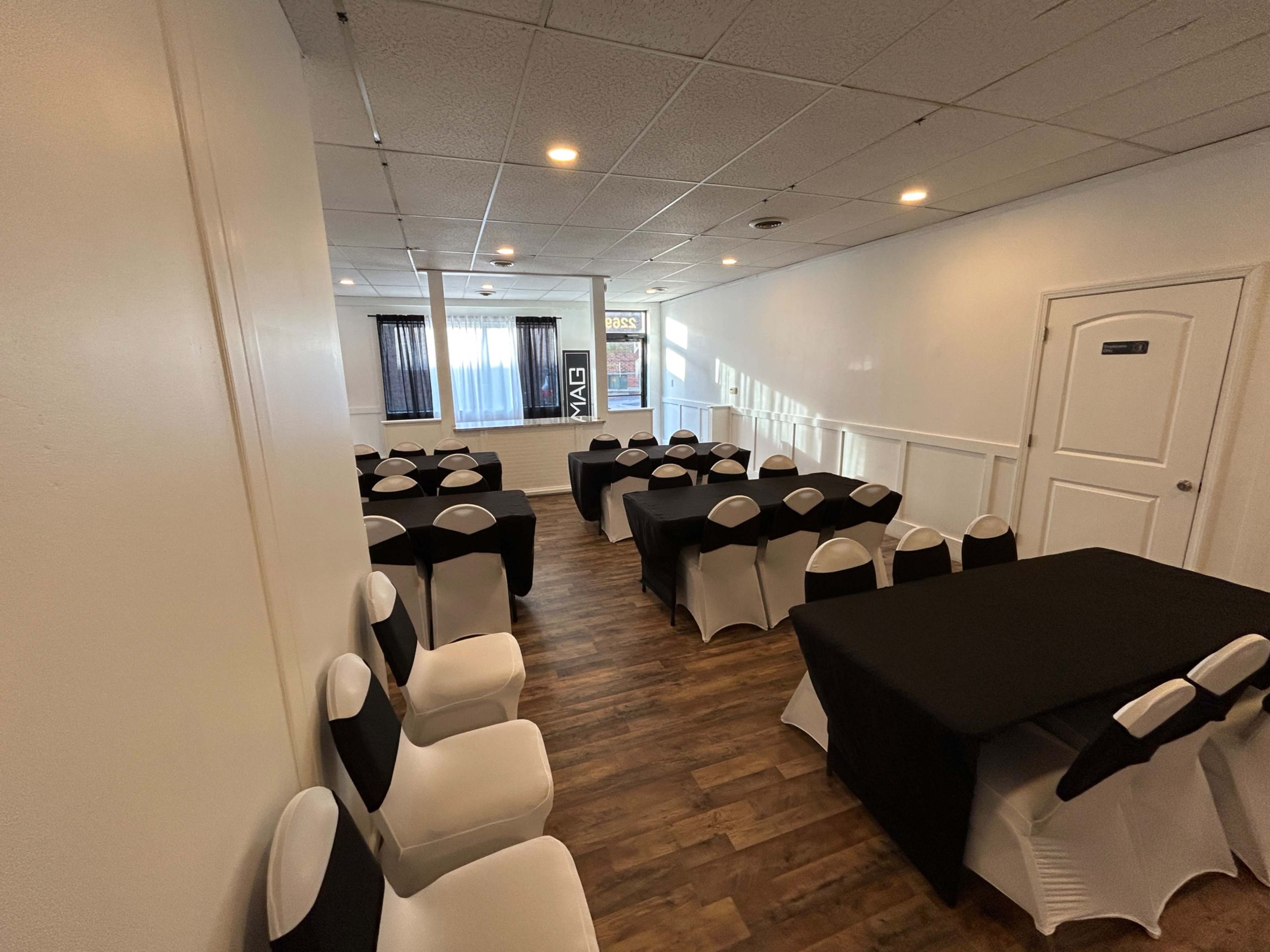 The image shows a well-lit event space set up with several tables covered in black cloth and adorned with white chair sashes.